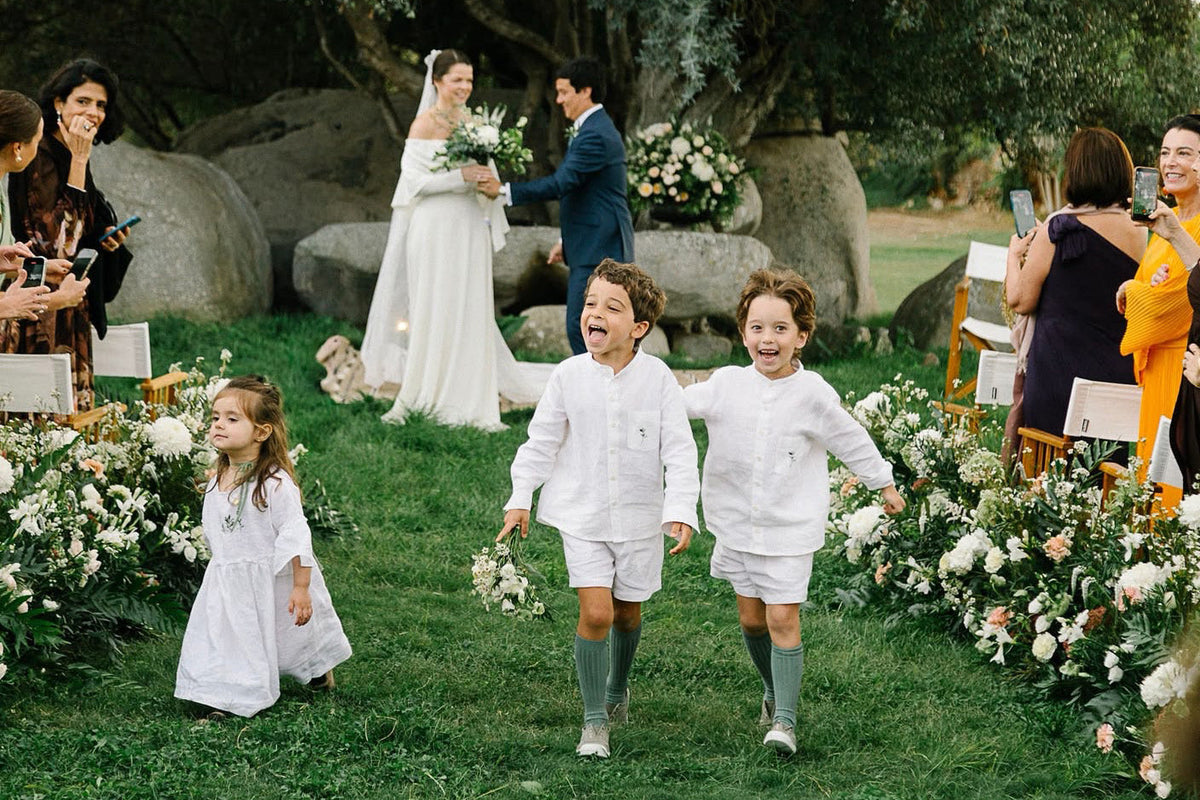 Pajens e daminha com peças de linho branco com com detalhe floral bordado, em cerimônia de casamento em jardim, durante o dia.