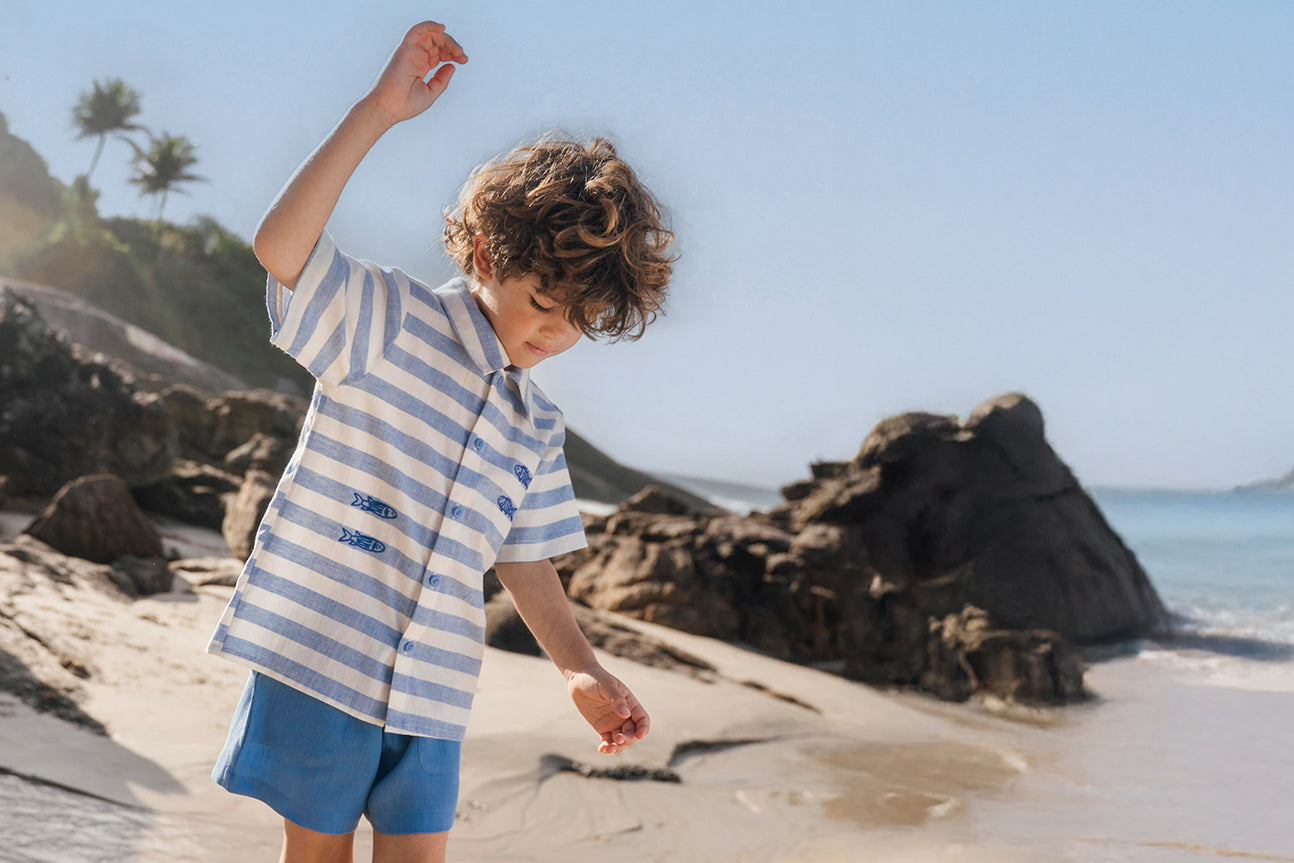 Menino se equilibrando em pedra na praia, vestindo camisa listrada bordada à mão com shorts de linho azul claro
