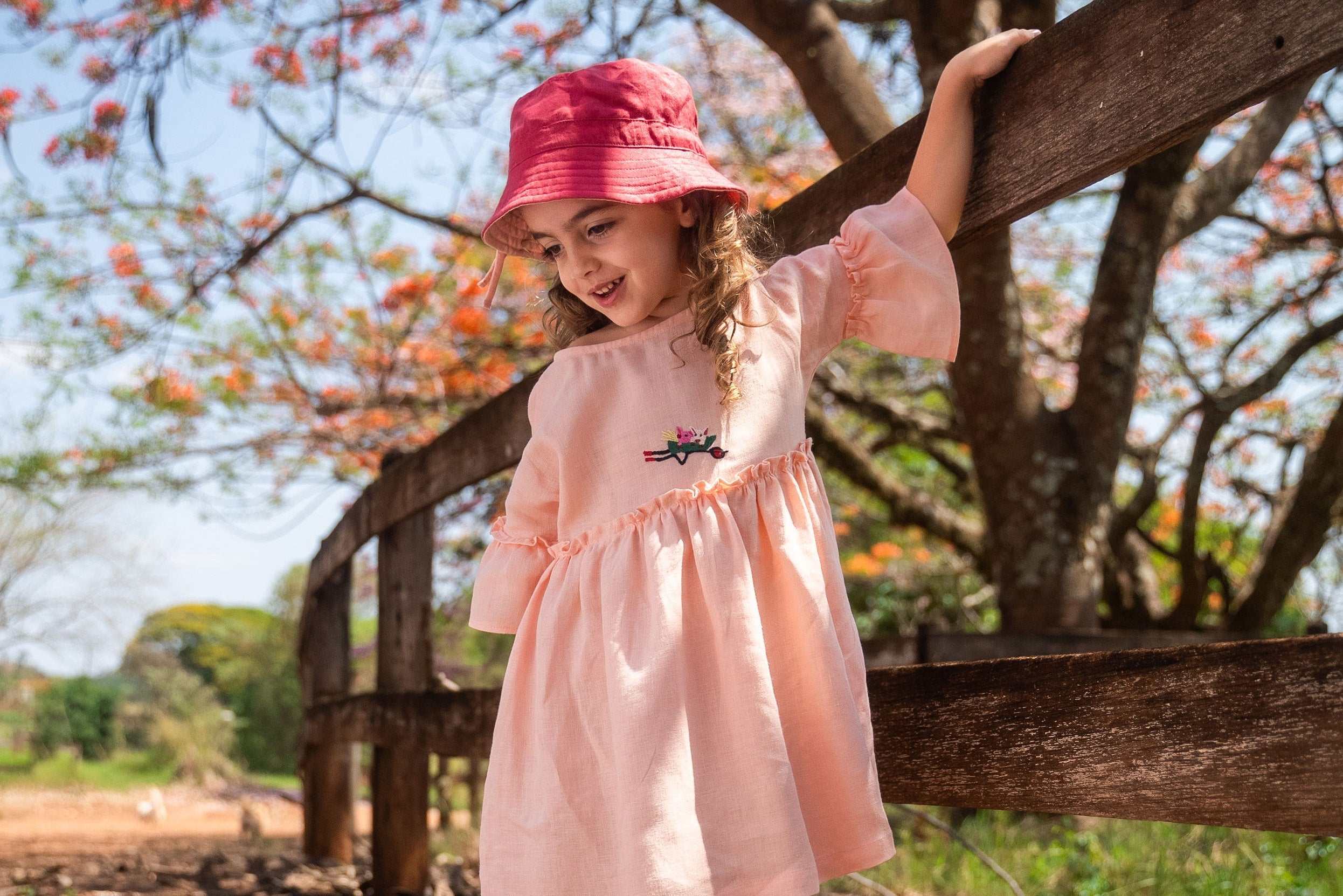 Menina em fazenda usando vestido rosa claro de linho puro com bordado à mão de porquinhos dentro de carrinho de mão, combinando com chapéu em sobre tom.