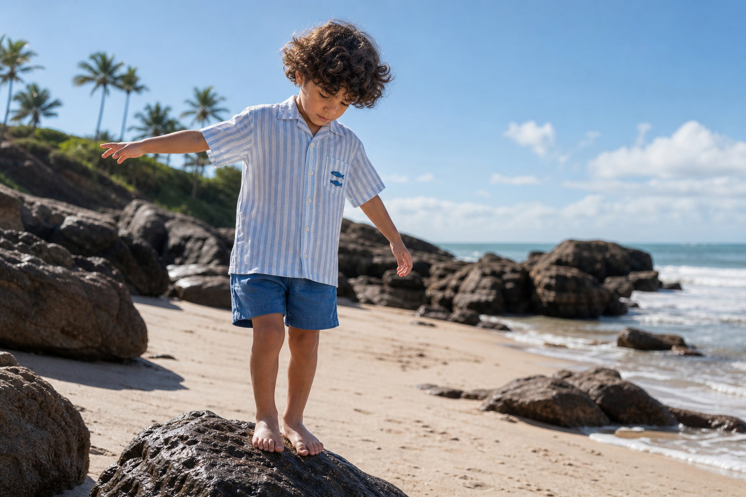 Menino de camisa de linho listrada azul e bermuda equilibrando-se em pedra na praia