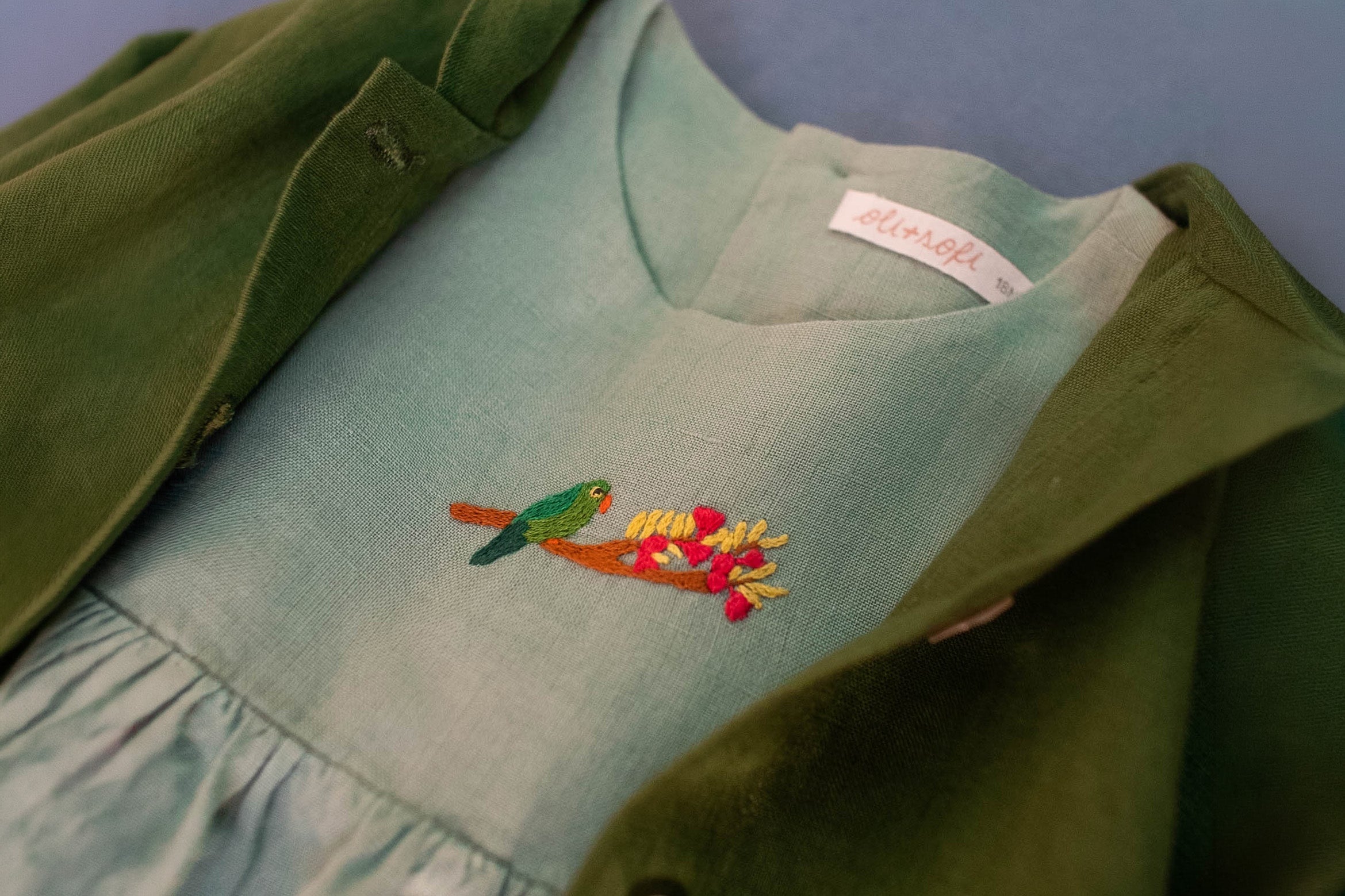 Vestido de linho puro azul visto em detalhe, com bordado de periquito no peito, coordenado com casaco verde também em linho puro