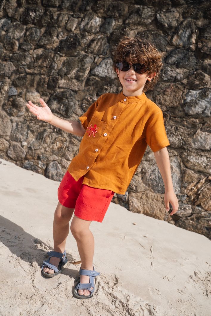 Menino na praia, vestindo shorts de linho vermelho e camisa terracota bom bordado de polvo em mesmo tom do shorts.