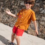 Menino na praia, vestindo shorts de linho vermelho e camisa terracota bom bordado de polvo em mesmo tom do shorts.