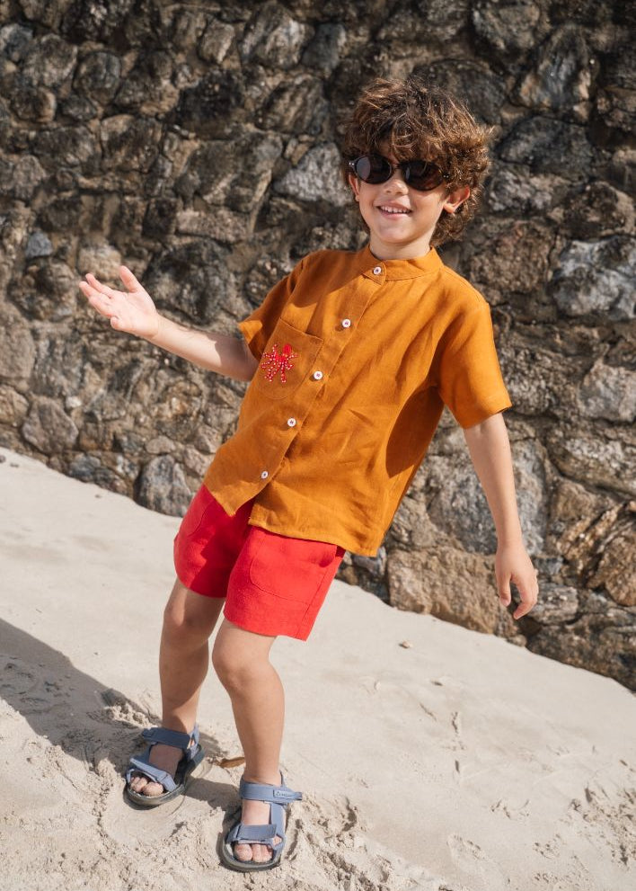 Menino na praia, vestindo shorts de linho vermelho e camisa terracota bom bordado de polvo em mesmo tom do shorts.