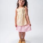 Menina vestindo Bata Flora infantil em algodão anarruga listrado amarelo com flores bordadas à mão e amarração no ombro, combinando com saia de linho listrada branco e rosa.