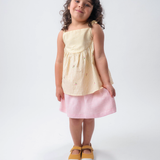 Menina vestindo Bata Flora infantil em algodão anarruga listrado amarelo com flores bordadas à mão e amarração no ombro, combinando com saia de linho listrada branco e rosa.