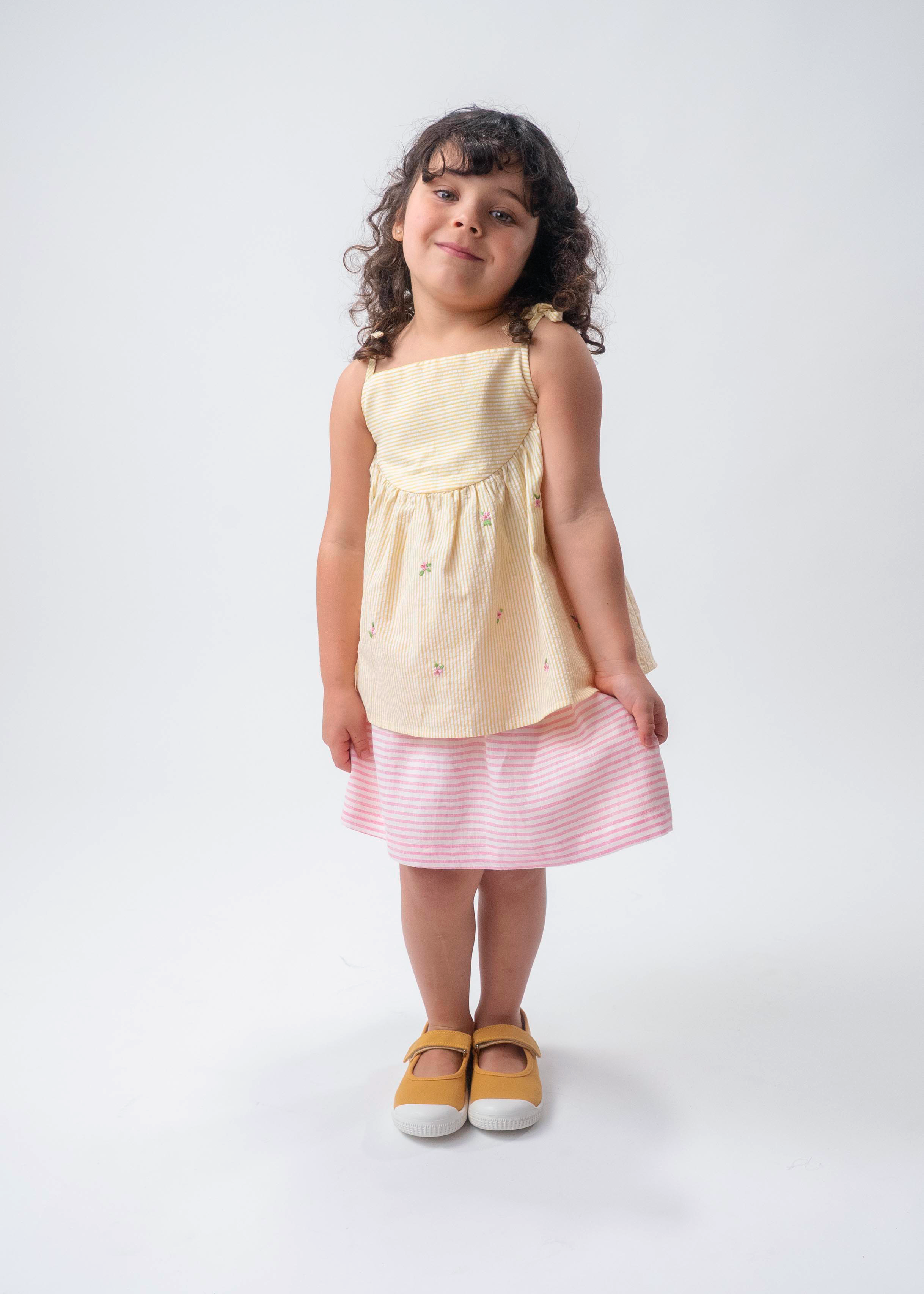 Menina vestindo Bata Flora infantil em algodão anarruga listrado amarelo com flores bordadas à mão e amarração no ombro, combinando com saia de linho listrada branco e rosa.