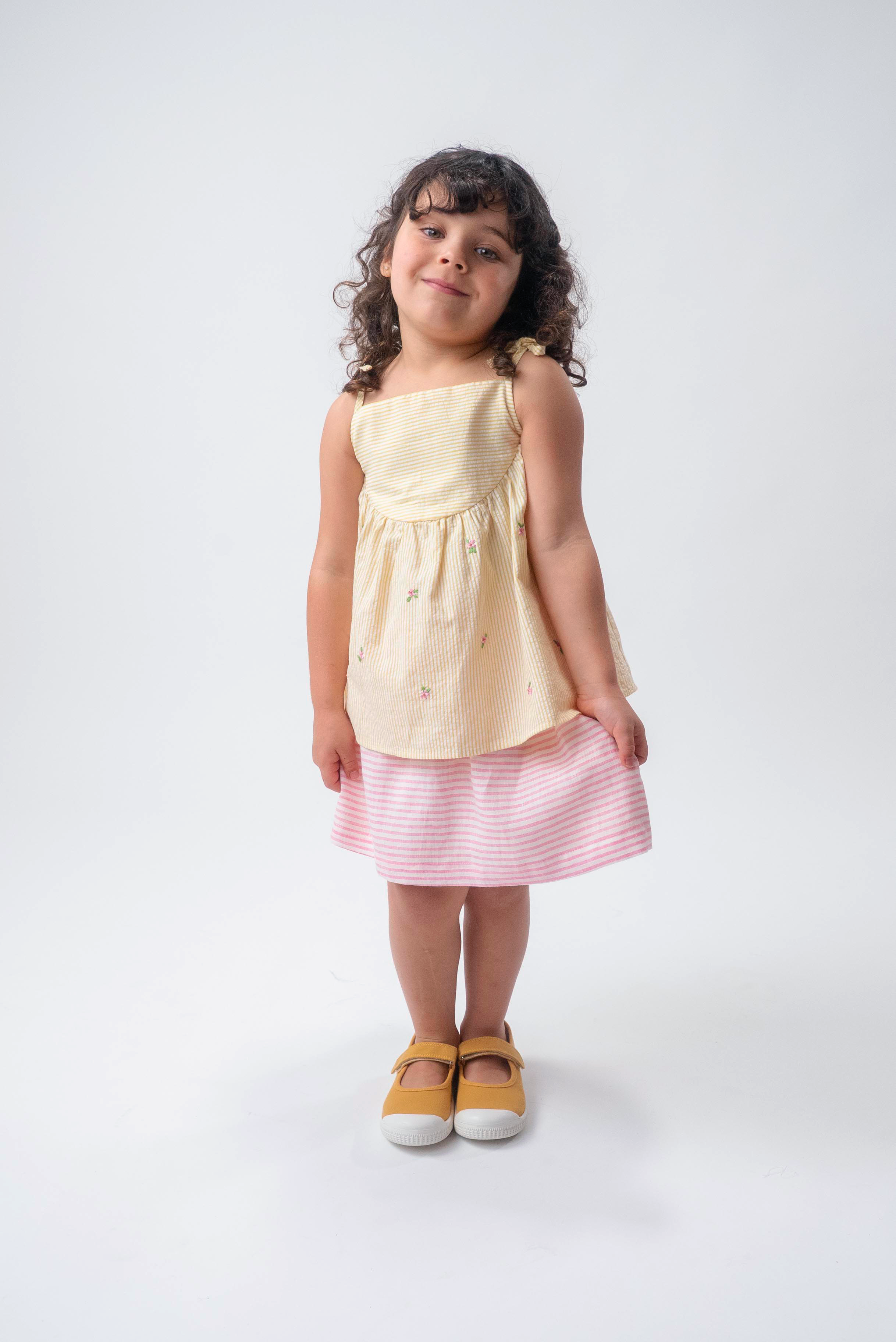 Menina vestindo Bata Flora infantil em algodão anarruga listrado amarelo com flores bordadas à mão e amarração no ombro, combinando com saia de linho listrada branco e rosa.