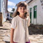 Menina vestindo bata infantil em algodão off-while com mangas curtas levemente bufantes, gola Peter Pan arredondada e pregas no peitilho.