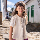 Menina vestindo bata infantil em algodão off-while com mangas curtas levemente bufantes, gola Peter Pan arredondada e pregas no peitilho.