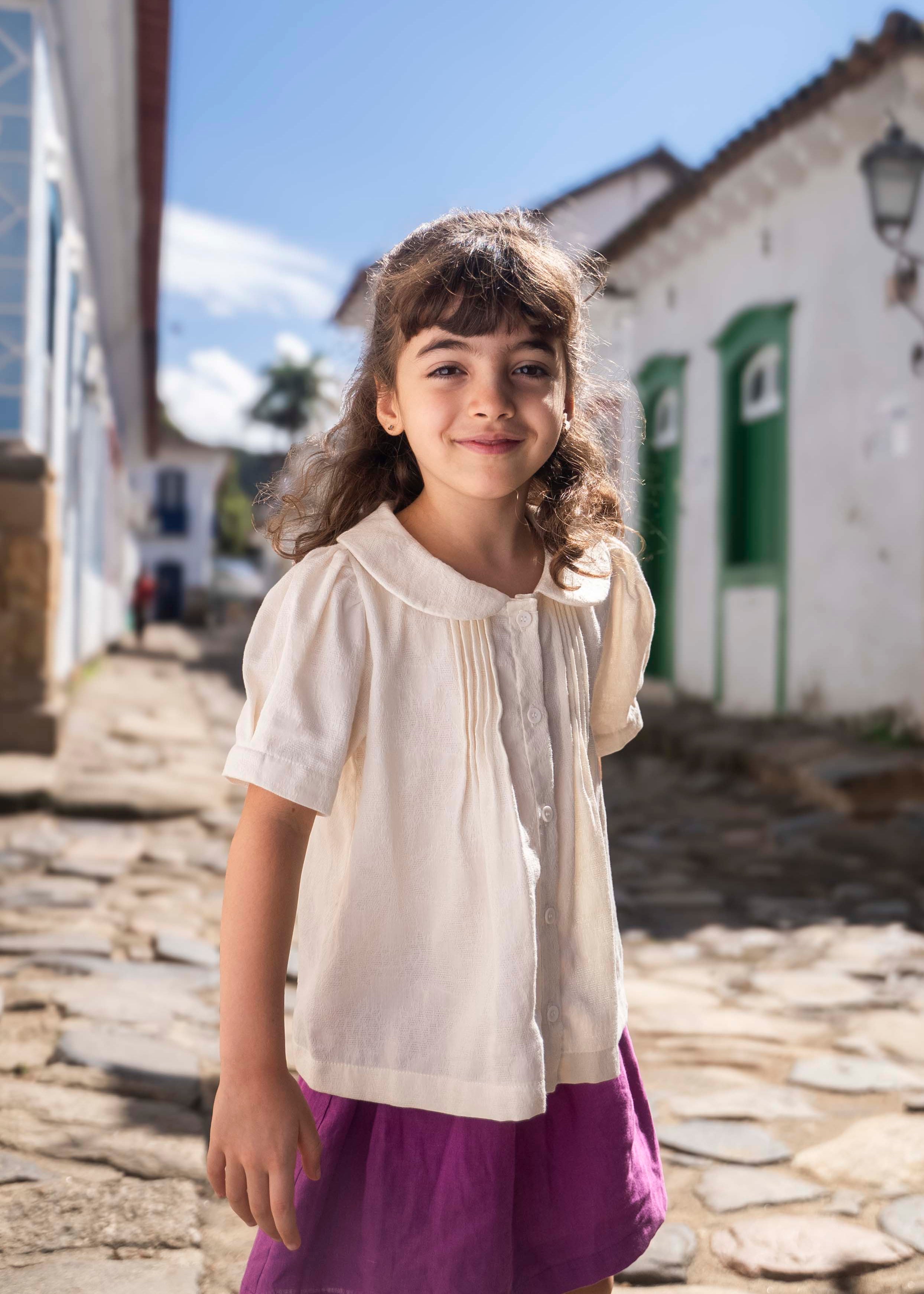 Menina vestindo bata infantil em algodão off-while com mangas curtas levemente bufantes, gola Peter Pan arredondada e pregas no peitilho.