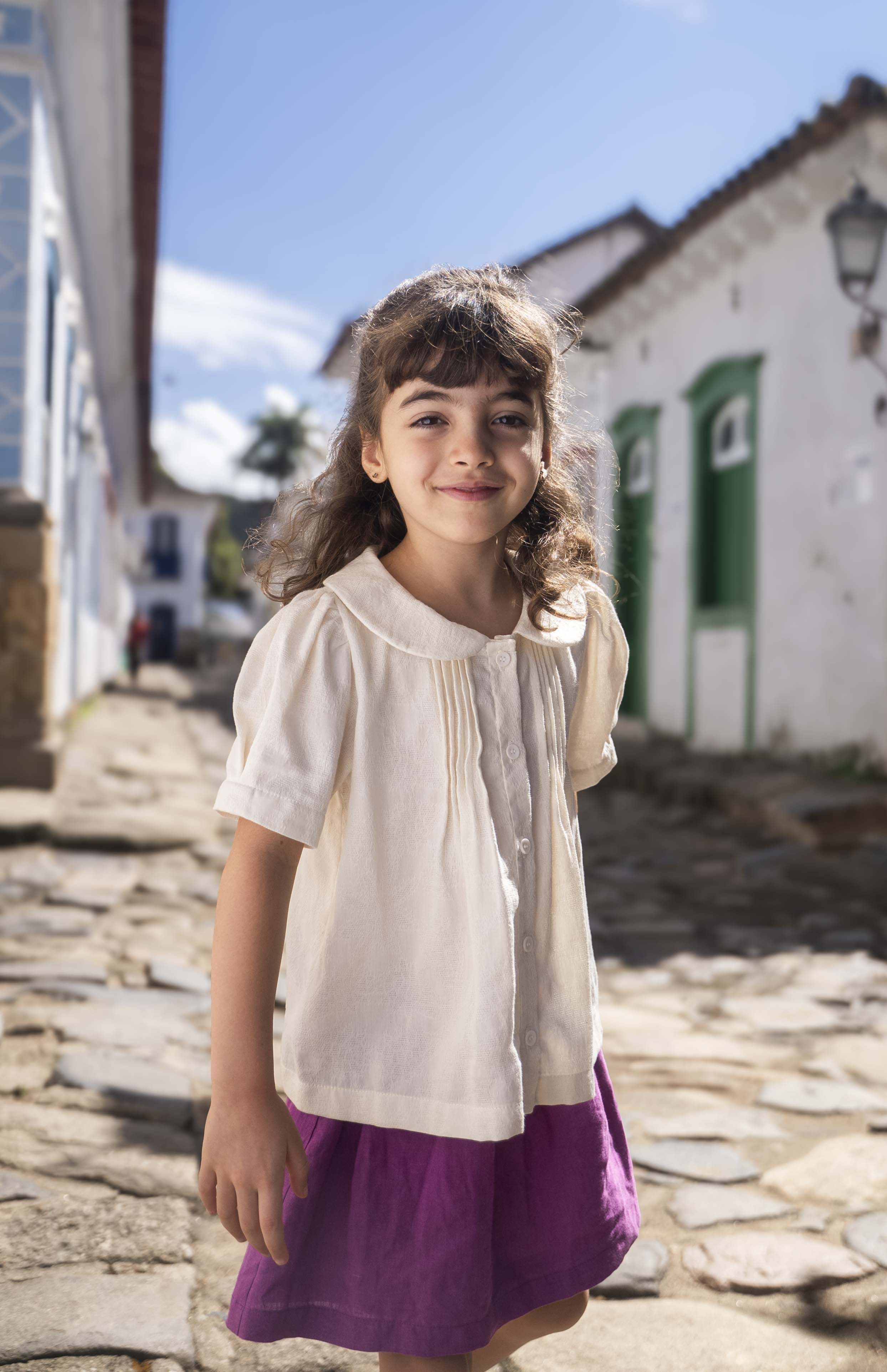 Menina vestindo bata infantil em algodão off-while com mangas curtas levemente bufantes, gola Peter Pan arredondada e pregas no peitilho.