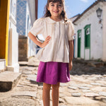 Menina vestindo bata infantil em algodão off-while com mangas curtas levemente bufantes, gola Peter Pan arredondada e pregas no peitilho.