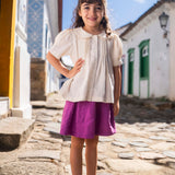 Menina vestindo bata infantil em algodão off-while com mangas curtas levemente bufantes, gola Peter Pan arredondada e pregas no peitilho.