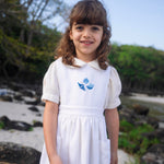 Menina vestindo bata infantil em algodão off-while com mangas curtas levemente bufantes e gola Peter Pan arredondada, em sobreposição com vestido branco.