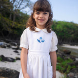 Menina vestindo bata infantil em algodão off-while com mangas curtas levemente bufantes e gola Peter Pan arredondada, em sobreposição com vestido branco.