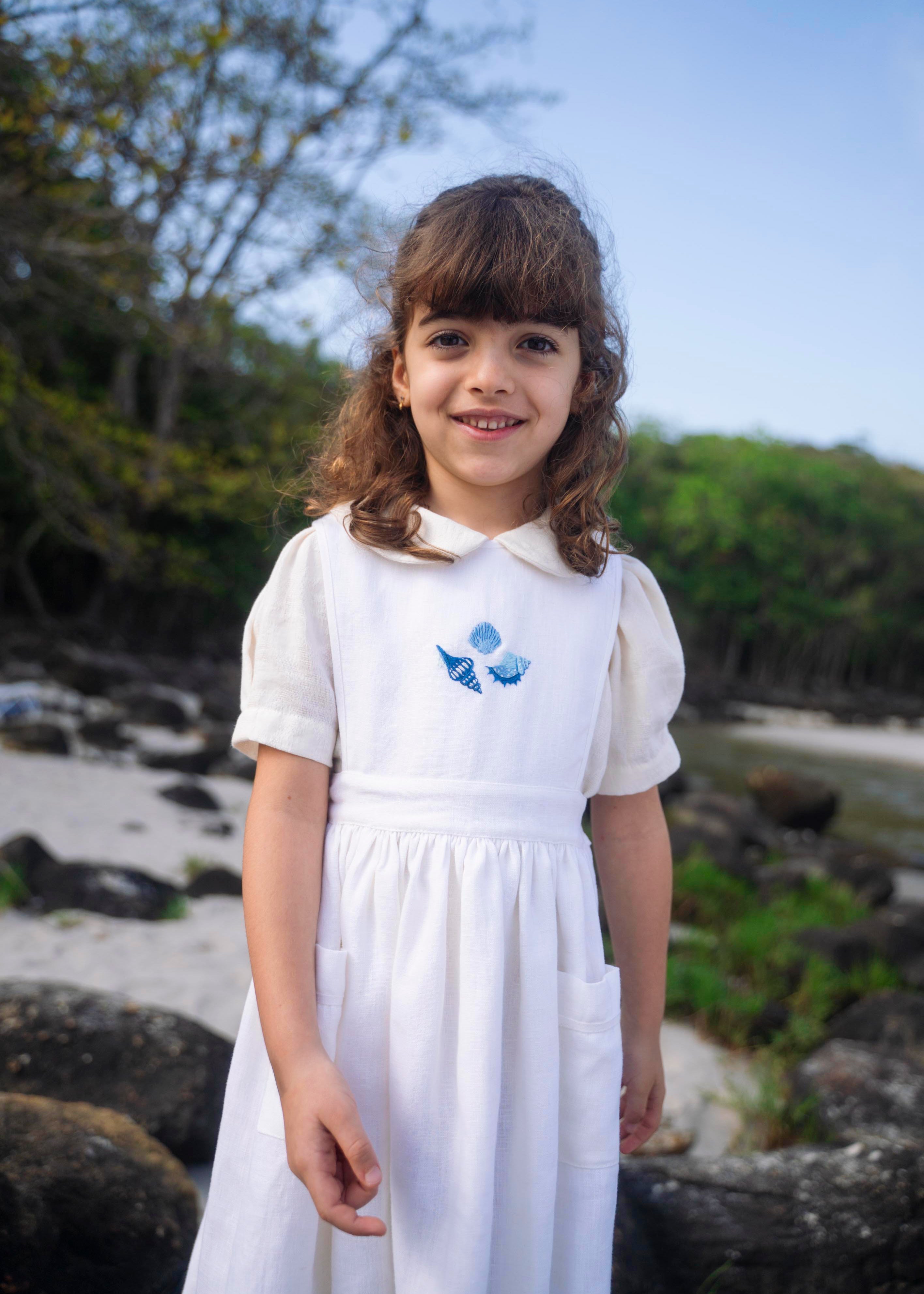 Menina vestindo bata infantil em algodão off-while com mangas curtas levemente bufantes e gola Peter Pan arredondada, em sobreposição com vestido branco.