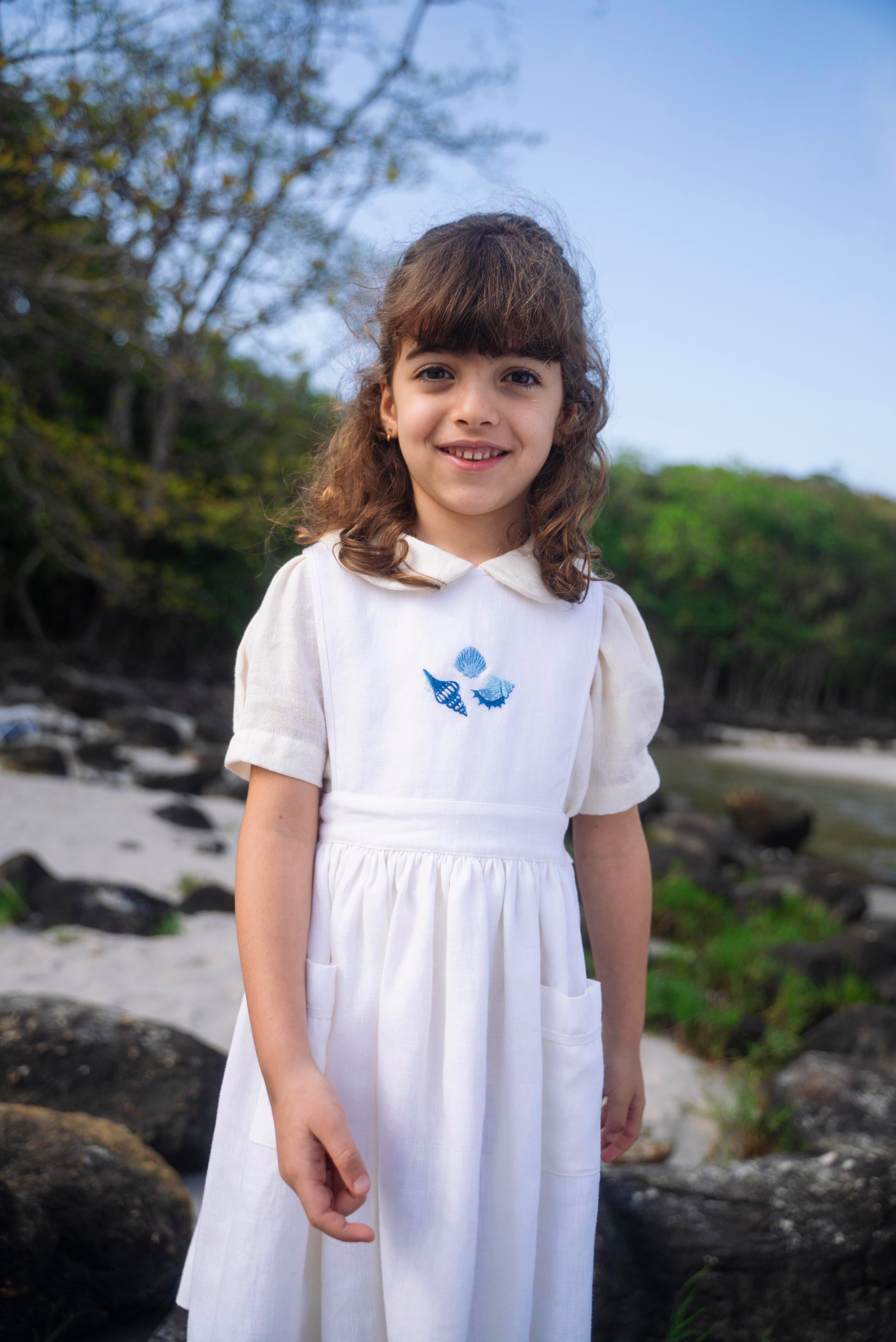 Menina vestindo bata infantil em algodão off-while com mangas curtas levemente bufantes e gola Peter Pan arredondada, em sobreposição com vestido branco.