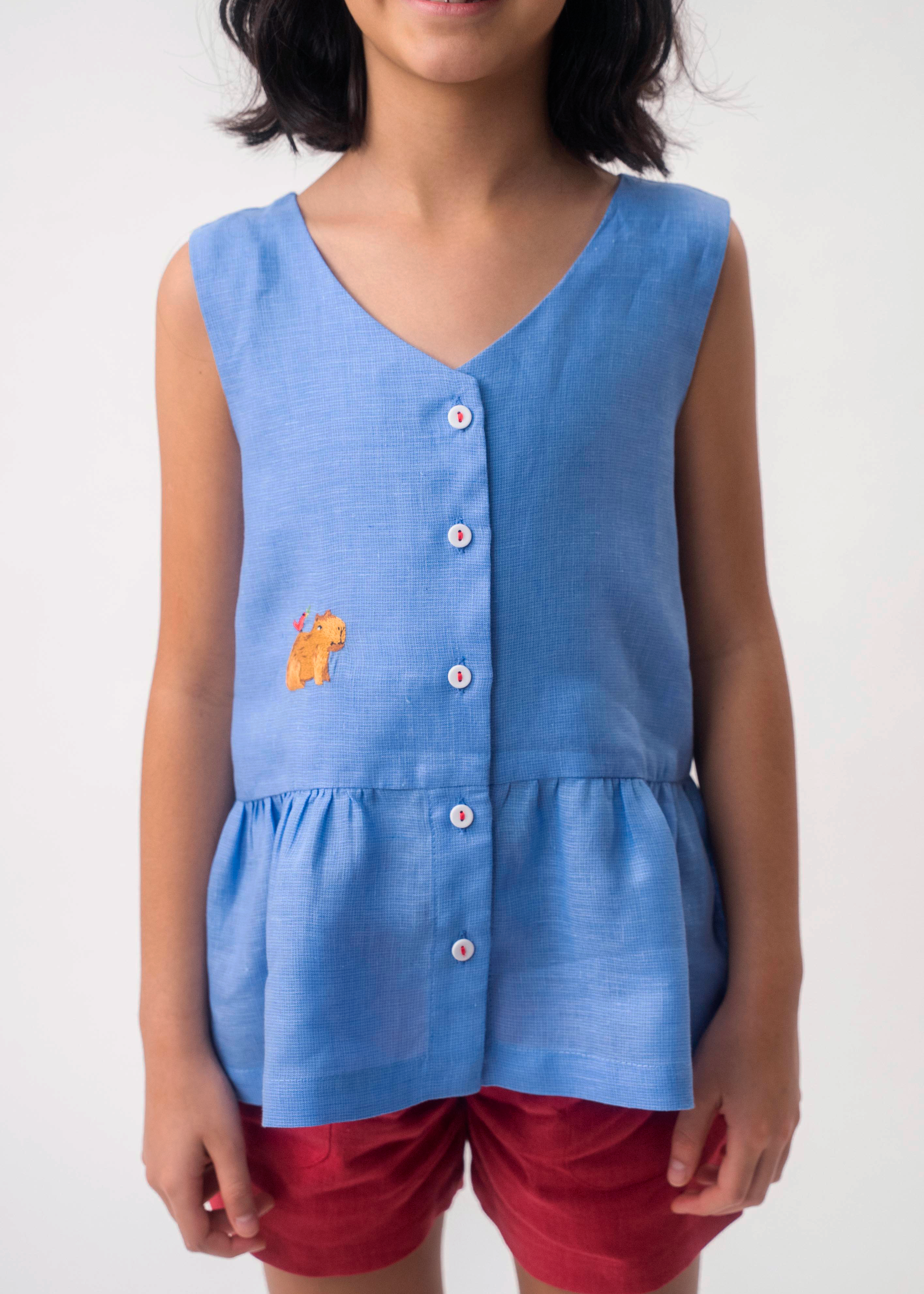 Menina vestindo Bata Lola infantil azul em 100% linho com decote V, botões frontais e bordado à mão, combinando com shorts vinho