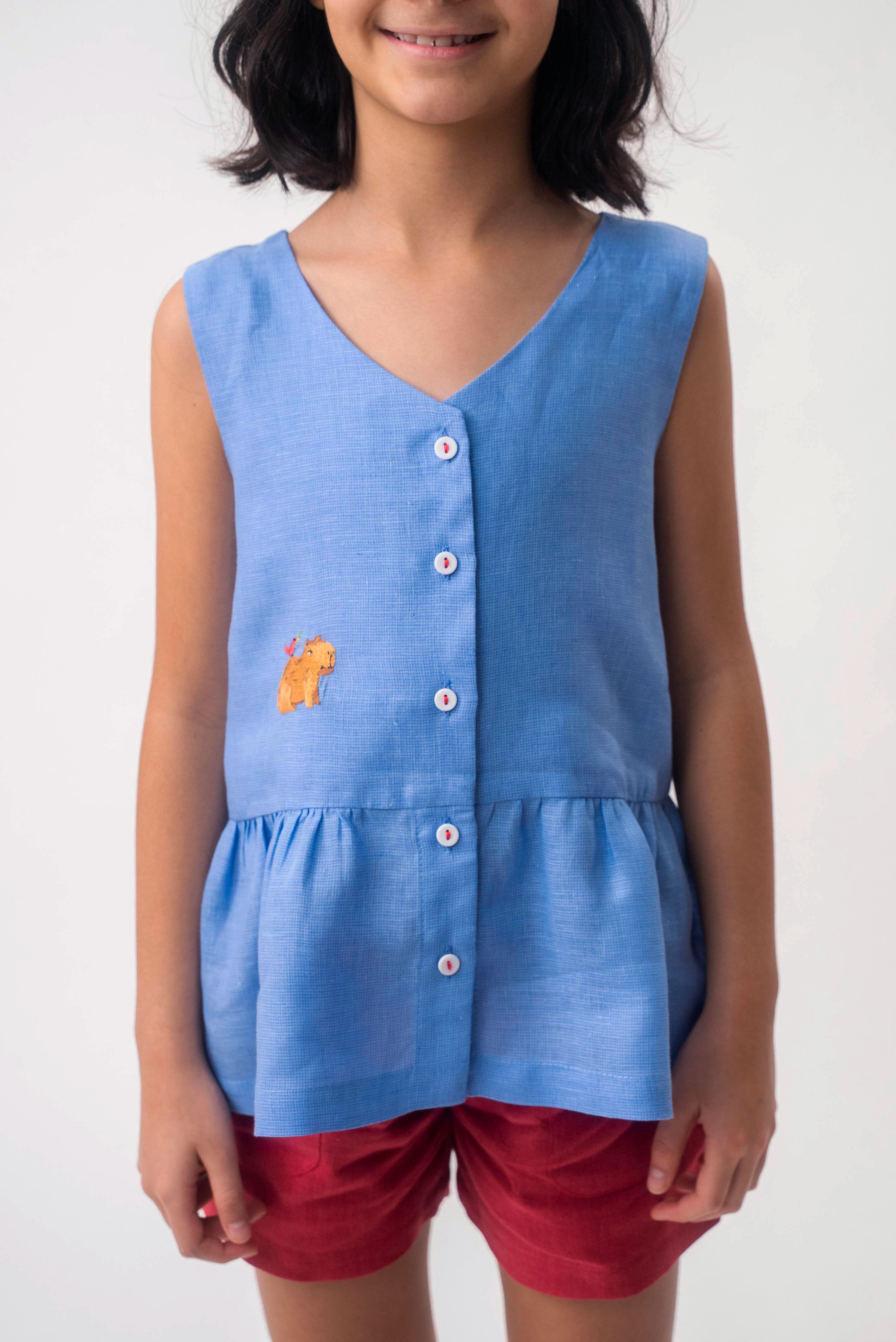 Menina vestindo Bata Lola infantil azul em 100% linho com decote V, botões frontais e bordado à mão, combinando com shorts vinho