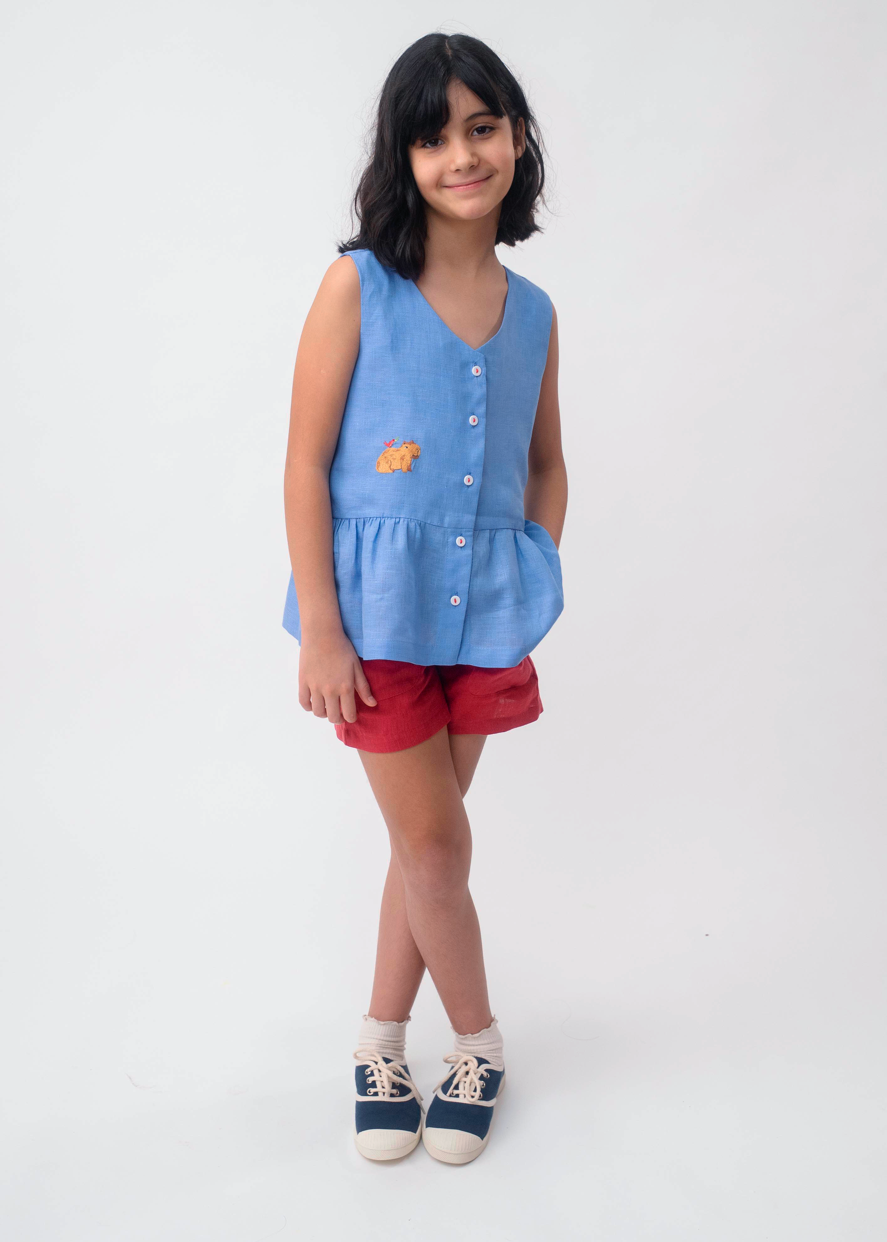Menina usando Bata Lola infantil em linho azul com bordado de capivara e shorts vermelho