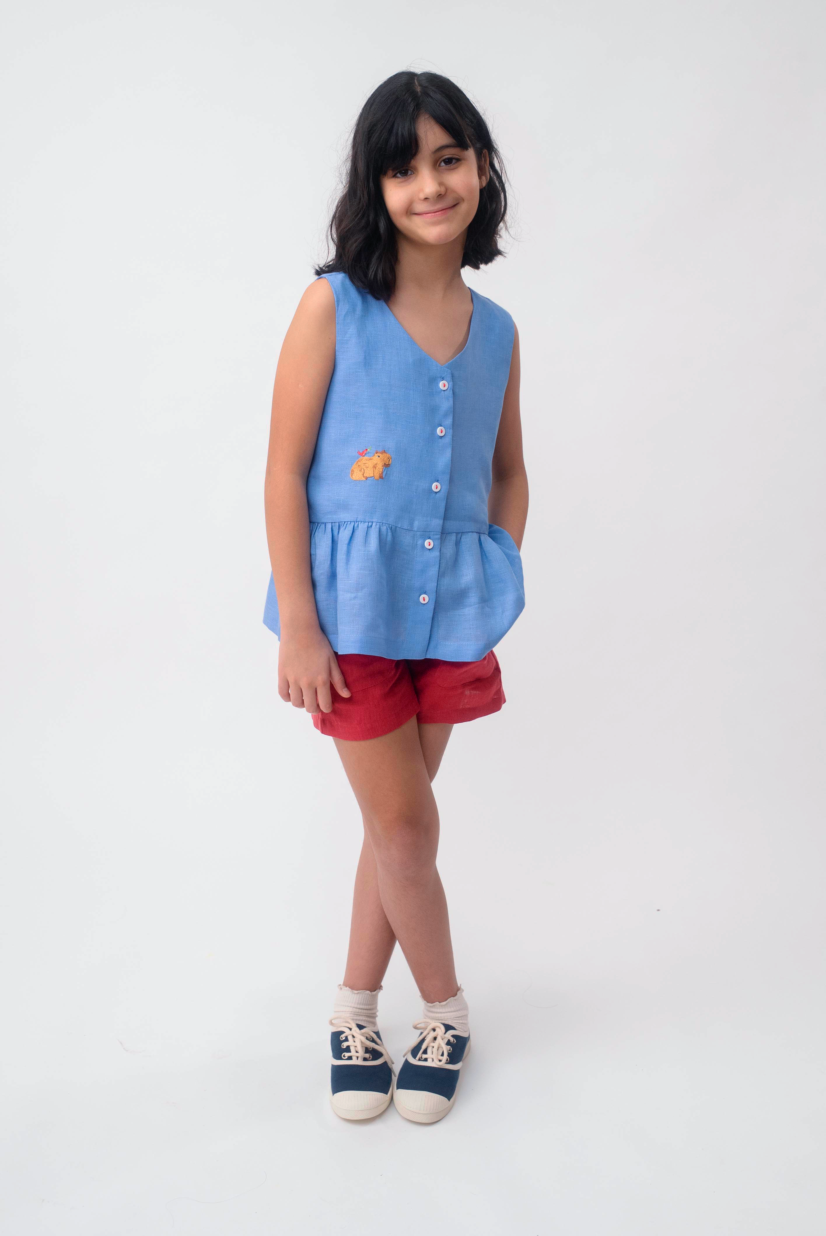 Menina usando Bata Lola infantil em linho azul com bordado de capivara e shorts vermelho