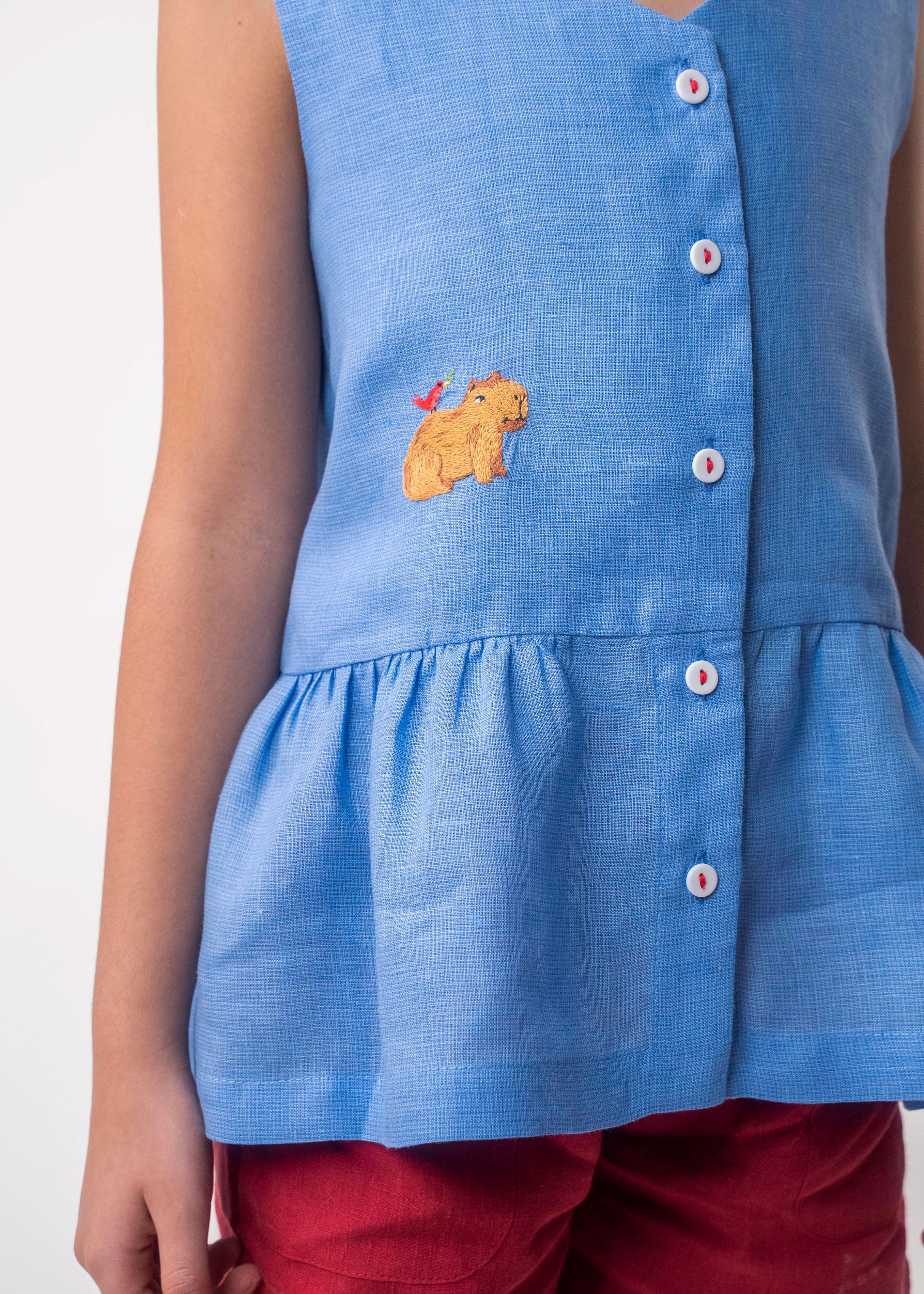 Detalhe de menina usando Bata Lola infantil em linho azul com bordado de capivara e barra franzida