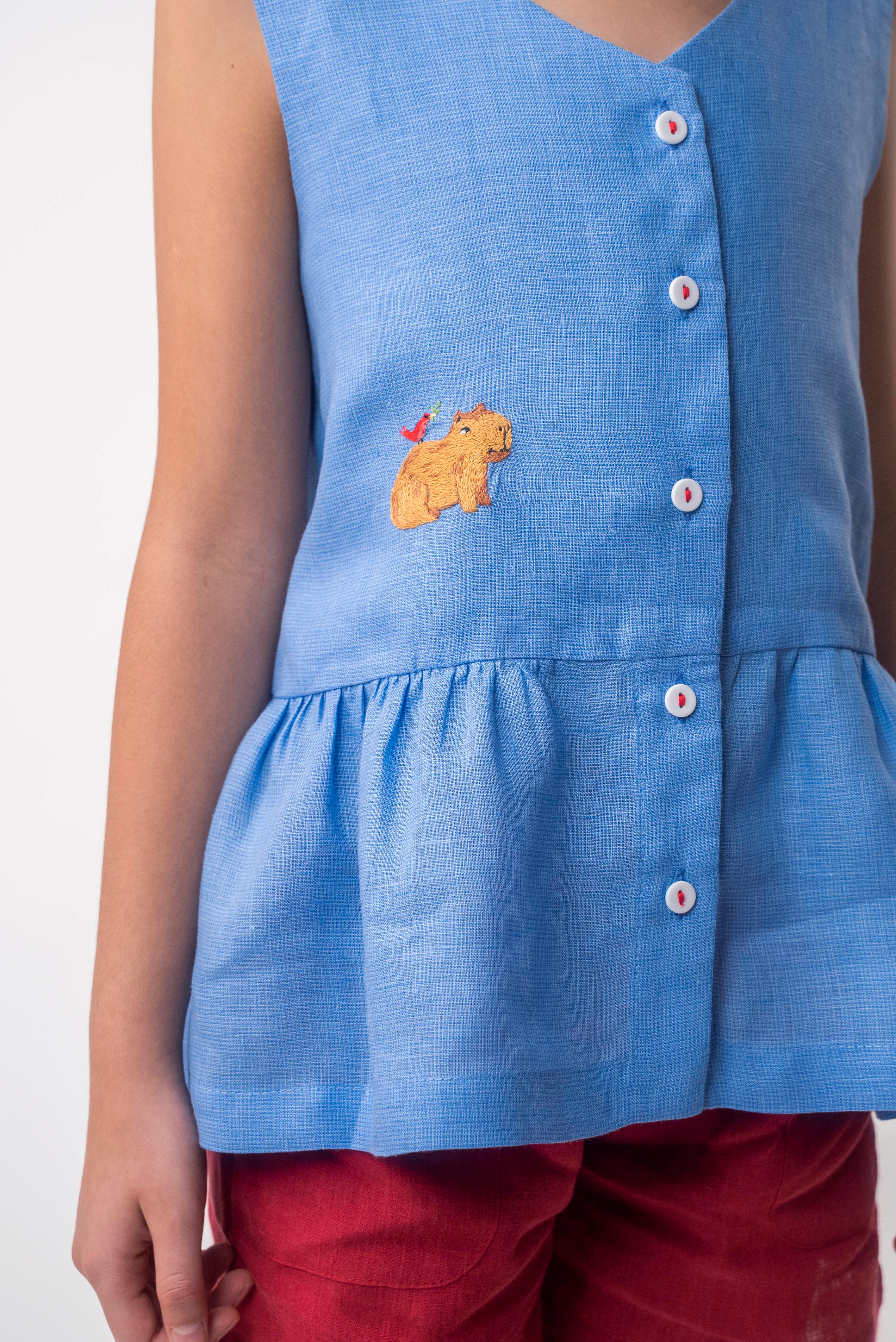 Detalhe de menina usando Bata Lola infantil em linho azul com bordado de capivara e barra franzida
