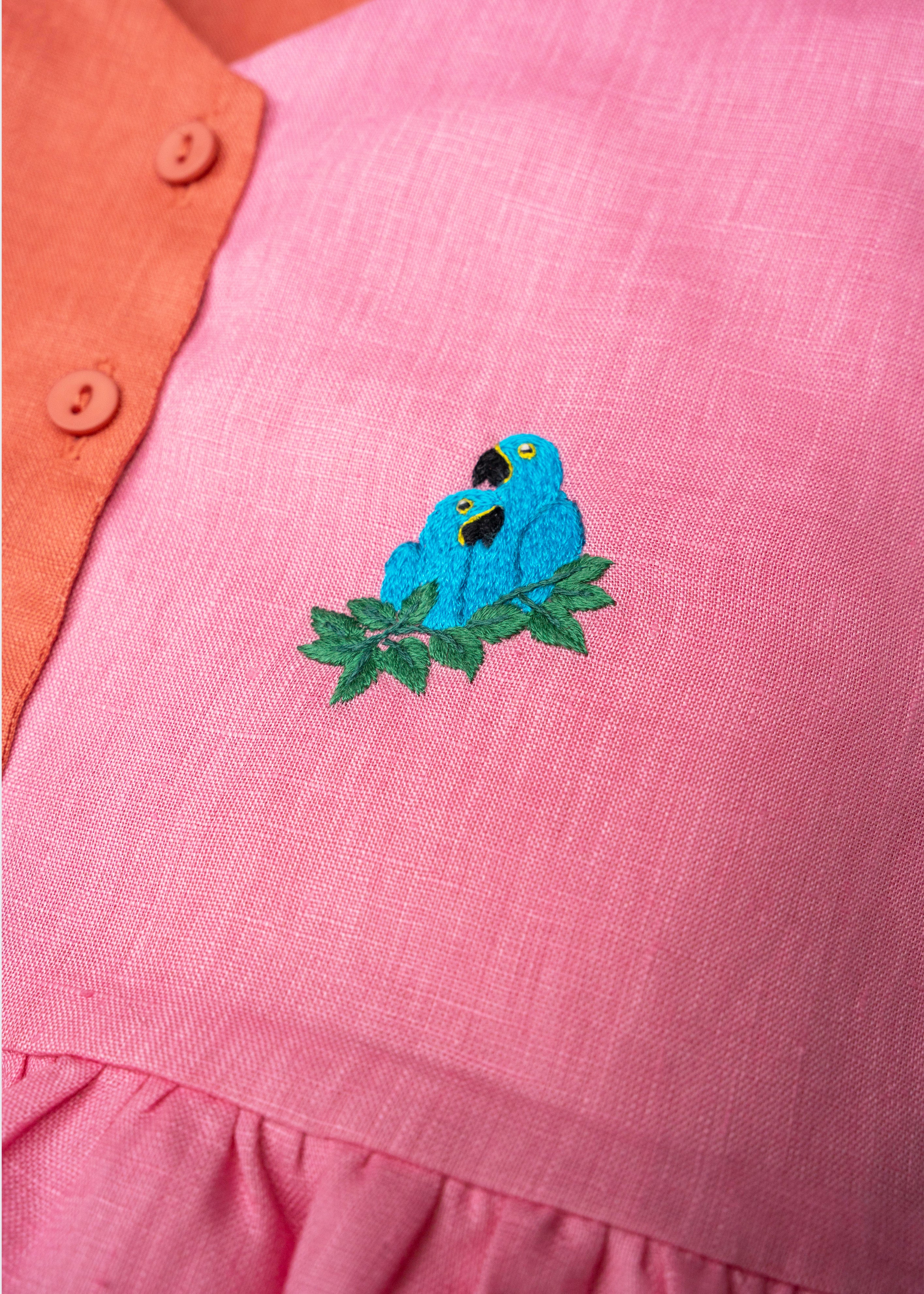 Detalhe da Bata Lola em linho rosa e coral com bordado à mão de duas araras-azuis entre folhas.
