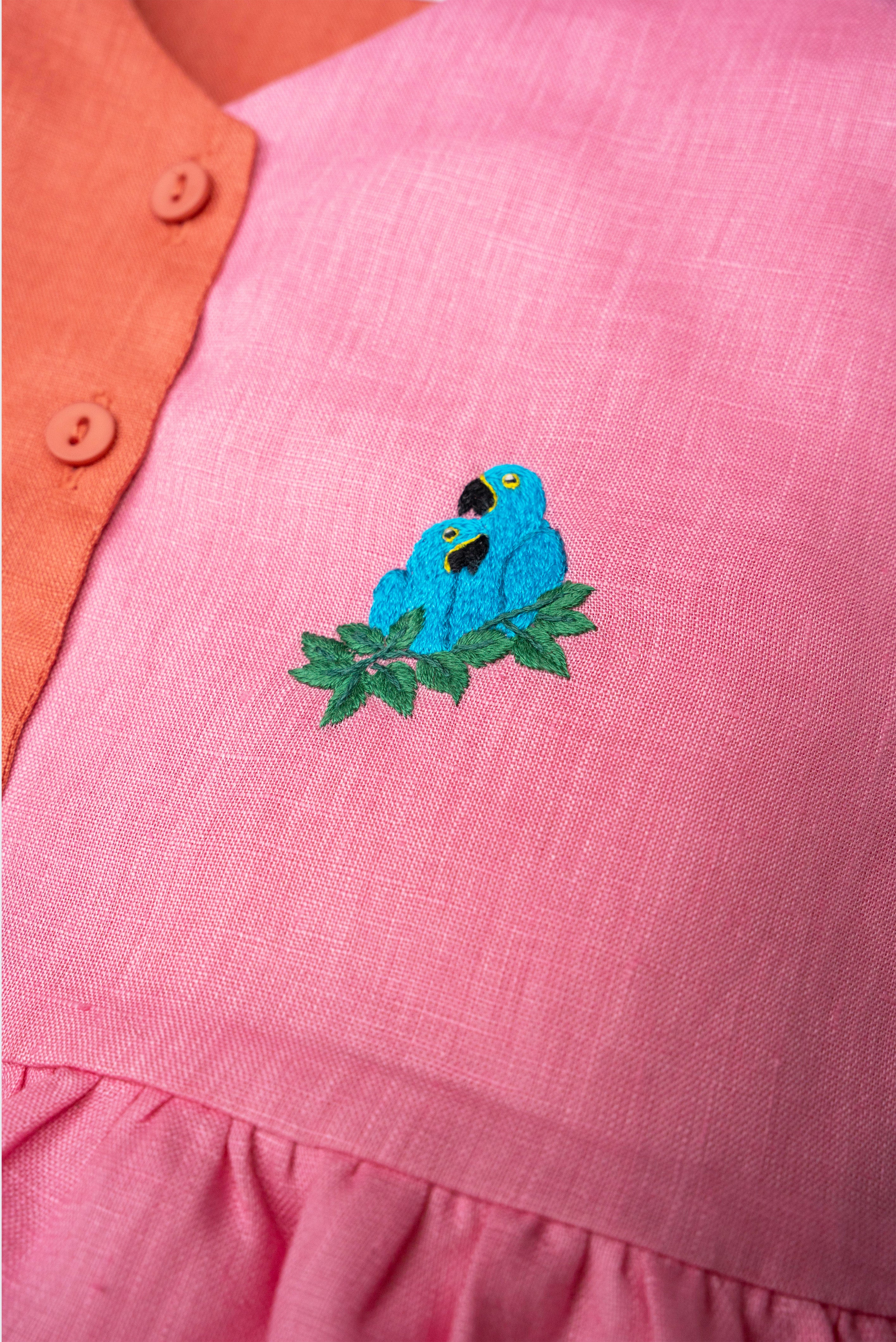 Detalhe da Bata Lola em linho rosa e coral com bordado à mão de duas araras-azuis entre folhas.