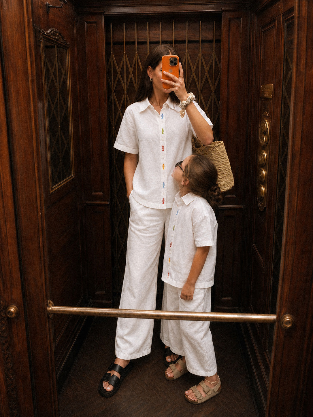 Mãe fazendo selfie em elevador com a filha vestindo camisa coordenada branca de linho com bordado de peixes coloridos entre botões da carcela, combinando com a menina.