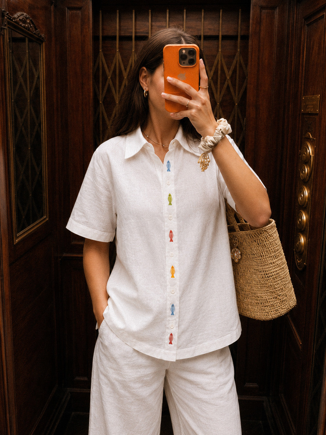 Mulher fazendo selfie em elevador vestindo camisa branca de linho com bordado de peixes coloridos entre botões da carcela.