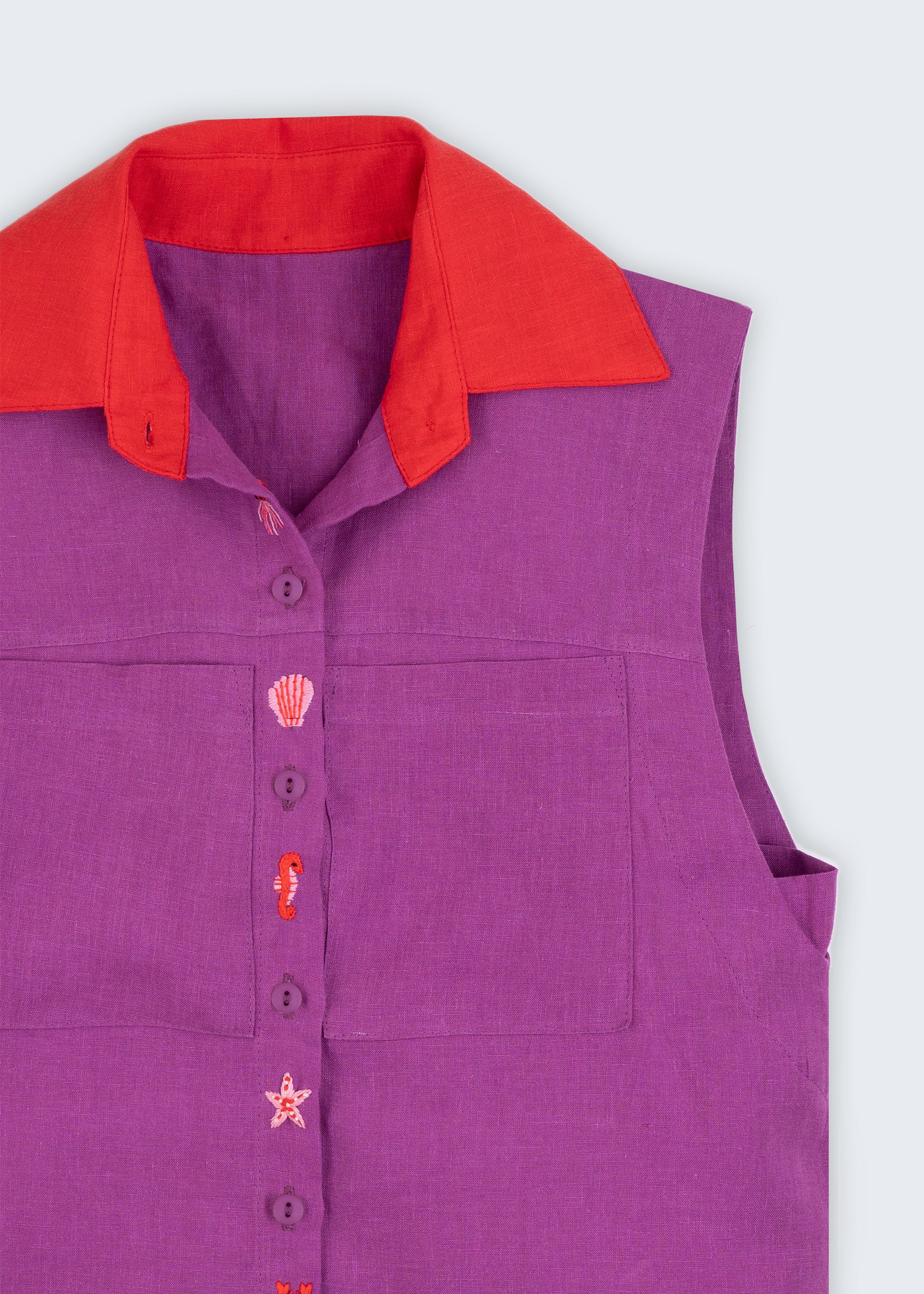 Camisa feminina sem mangas em linho roxo com gola vermelha e abotoamento bordado com conchas e cavalos-marinhos, vista em detalhe.
