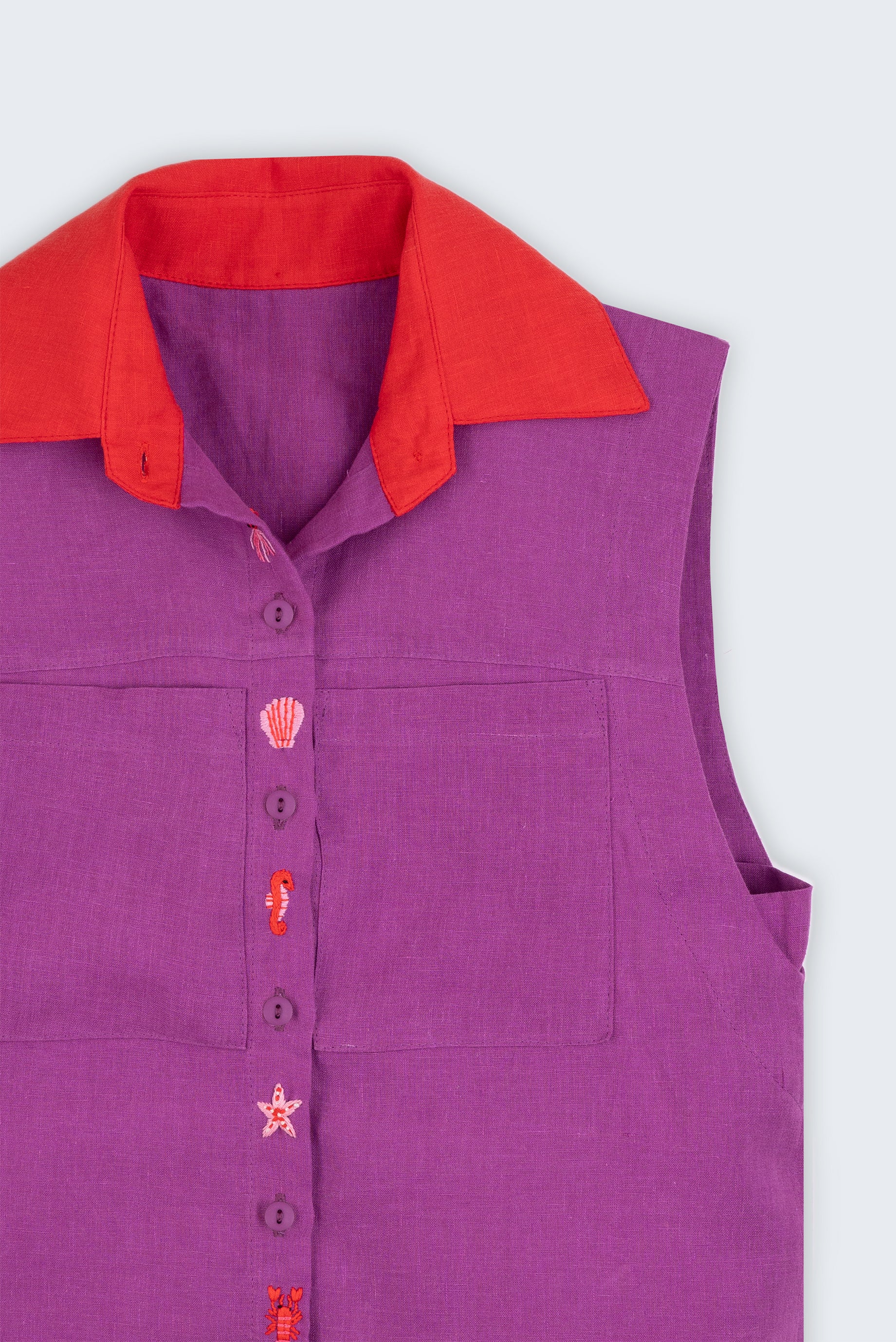 Camisa feminina sem mangas em linho roxo com gola vermelha e abotoamento bordado com conchas e cavalos-marinhos, vista em detalhe.