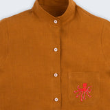 Camisa feminina adulto em linho puro terracota com gola padre, bolso bordado à mão com polvo vermelho e botões brancos, vista em detalhe.