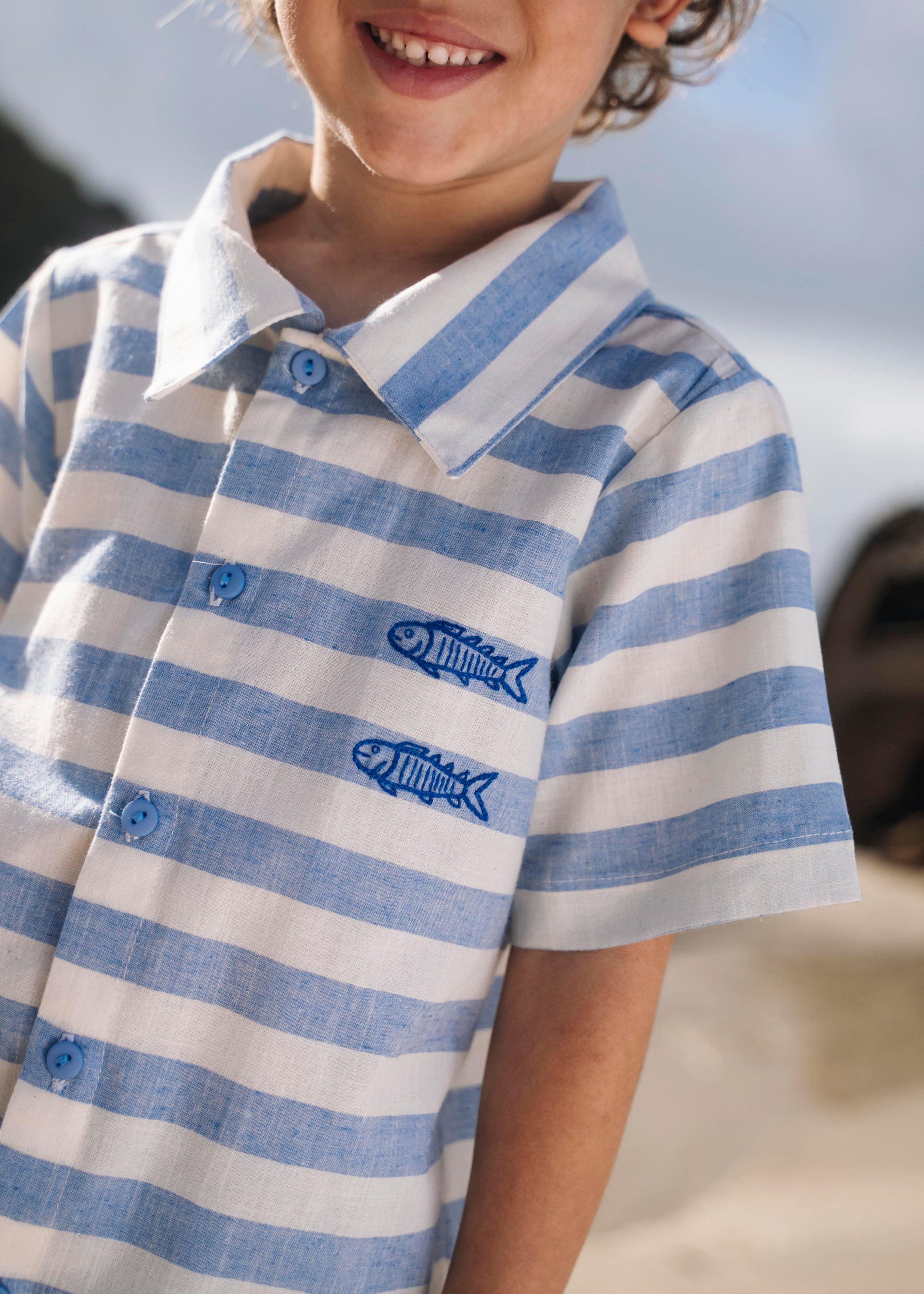 Close de menino com camisa infantil listrada azul em algodão com peixes bordados.