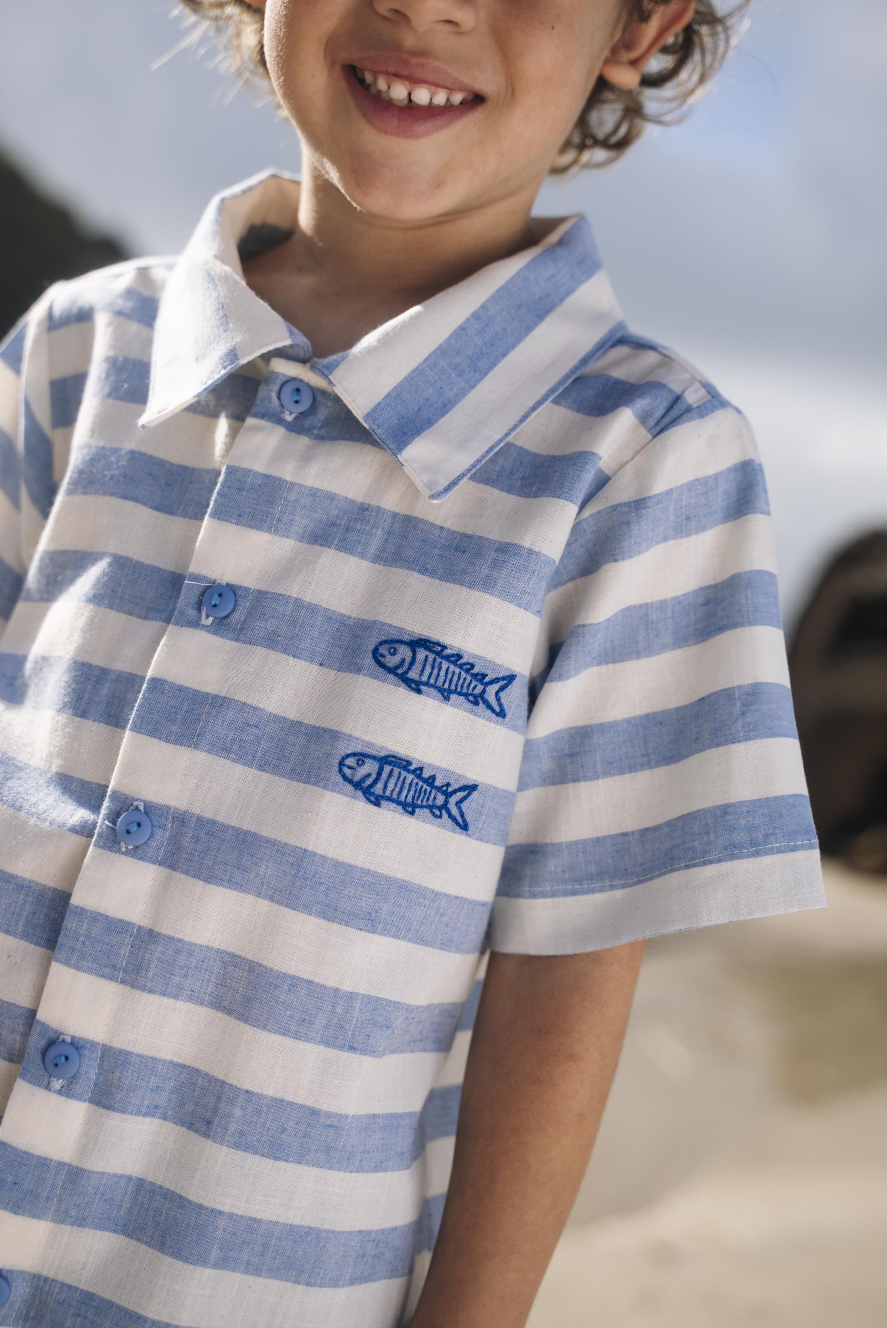 Close de menino com camisa infantil listrada azul em algodão com peixes bordados.