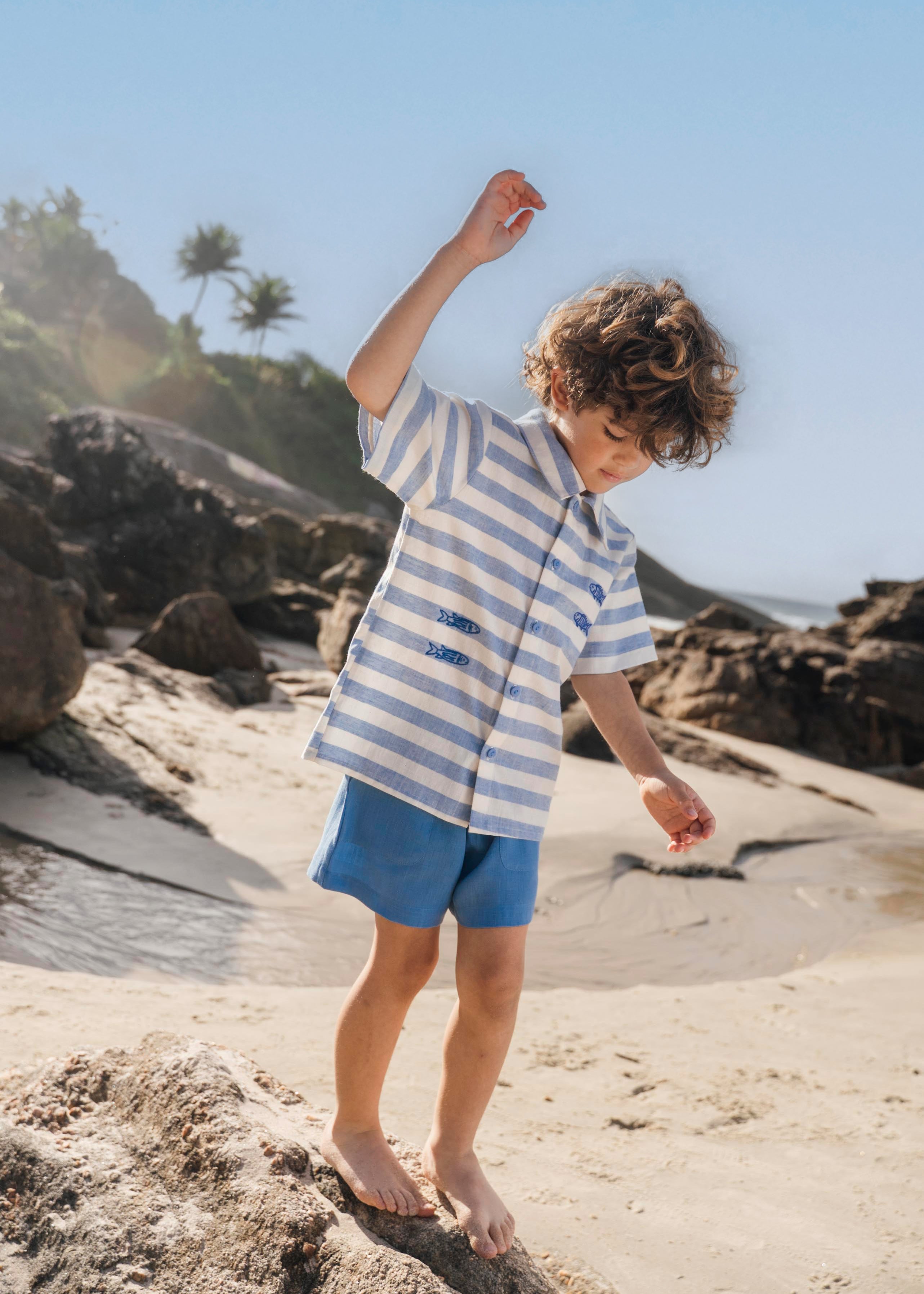 Menino com camisa infantil listrada azul em algodão BCI com peixes bordados e shorts azul combinando.