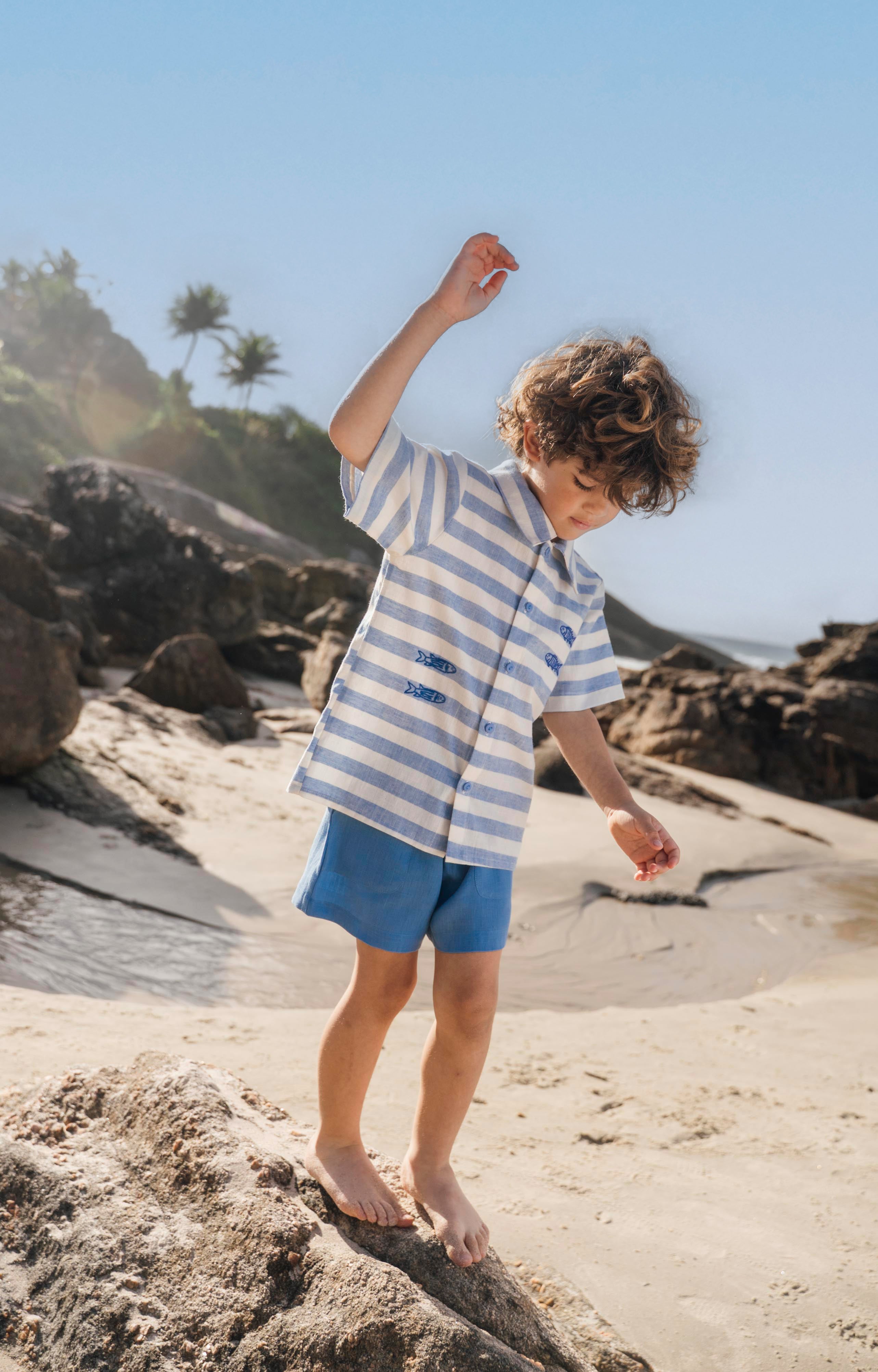 Menino com camisa infantil listrada azul em algodão BCI com peixes bordados e shorts azul combinando.