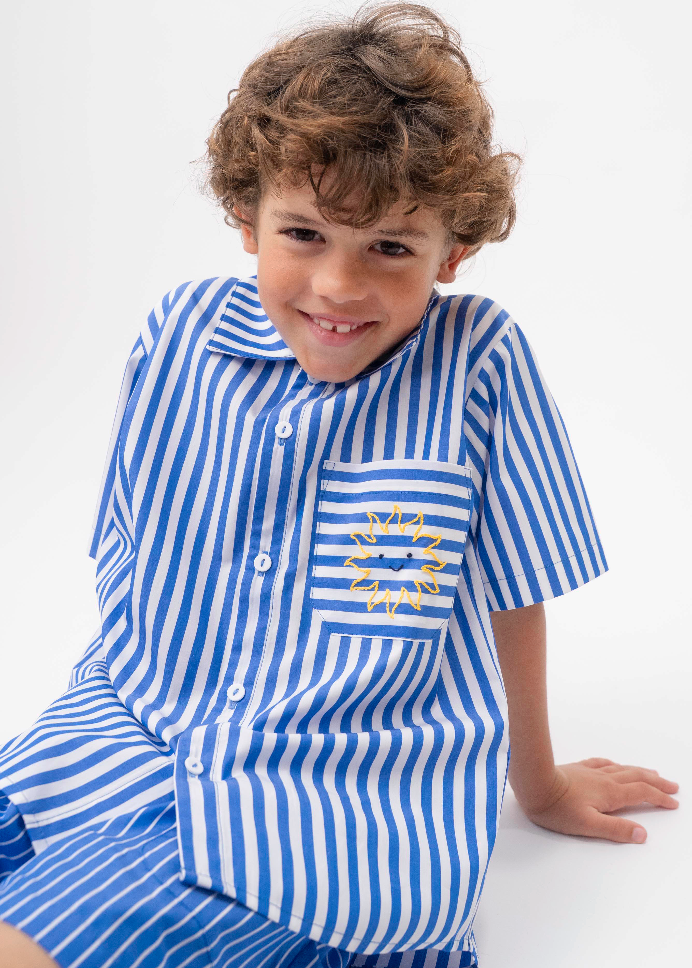 Detalhe da Camisa Gil listrada azul e branca vestida, com bolso de listras horizontais e bordado de sol amarelo.