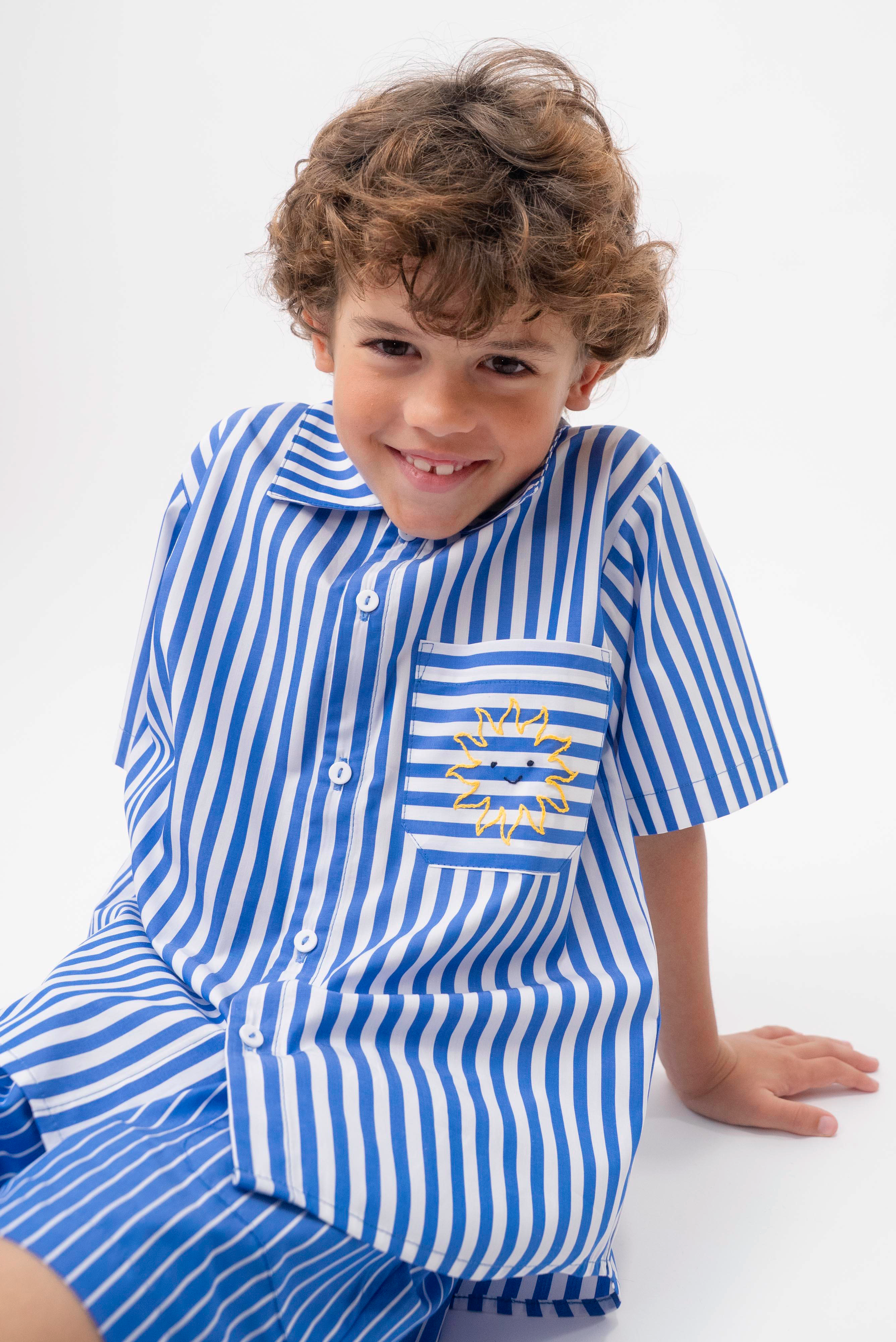 Detalhe da Camisa Gil listrada azul e branca vestida, com bolso de listras horizontais e bordado de sol amarelo.