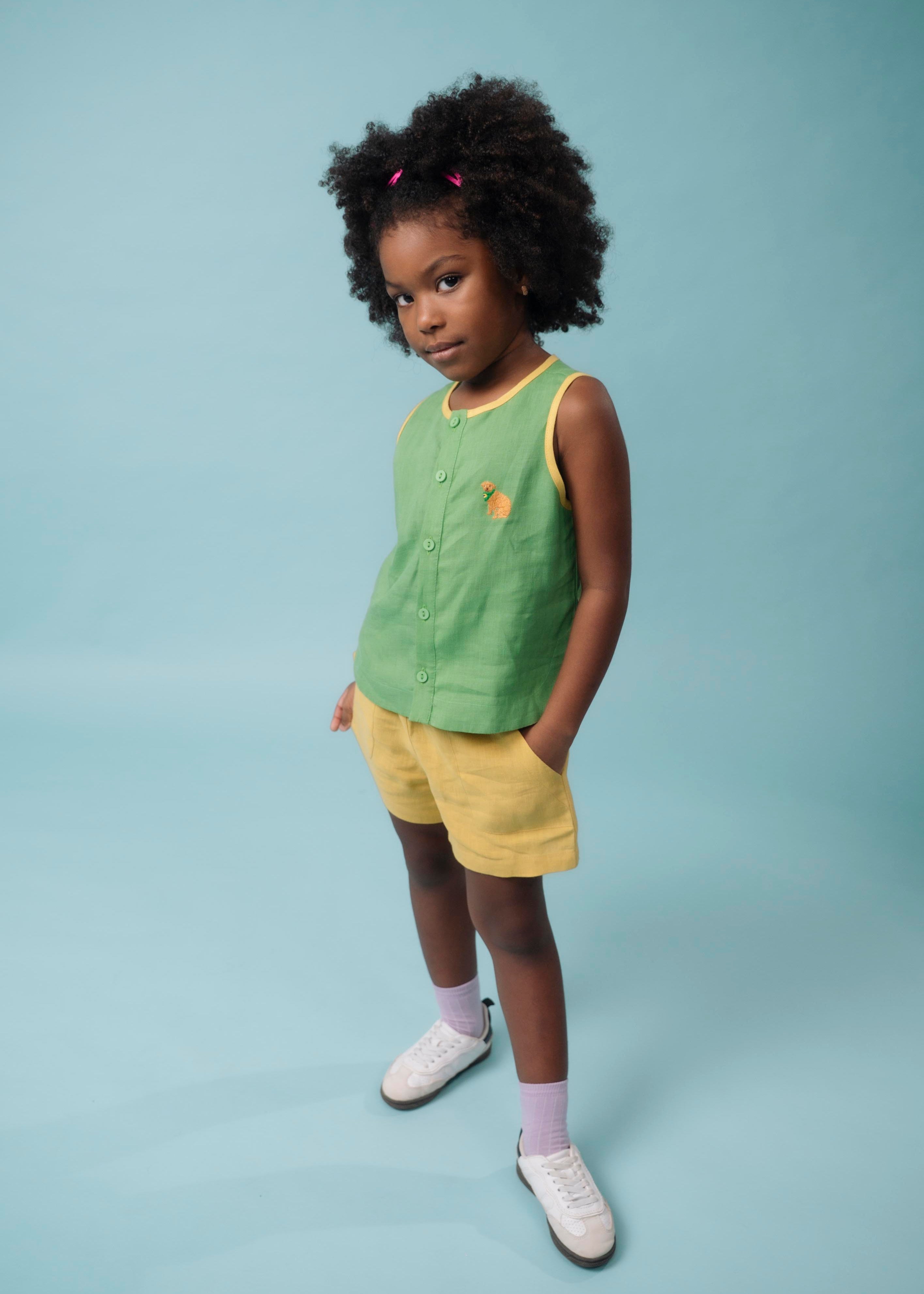 Menina usando camisa infantil em linho verde com acabamento amarelo e bordado à mão de cachorro caramelo, combinando com shorts amarelo
