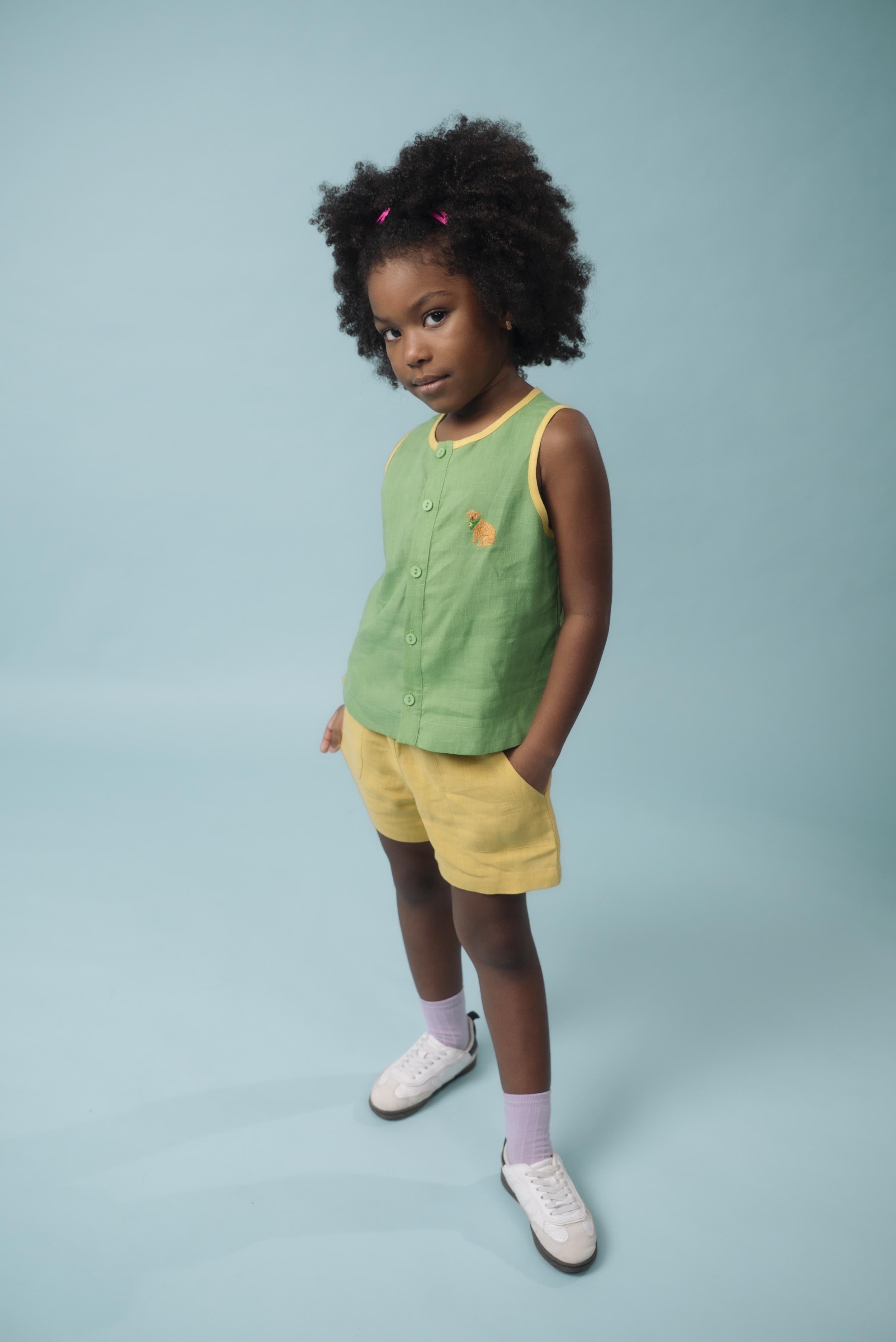 Menina usando camisa infantil em linho verde com acabamento amarelo e bordado à mão de cachorro caramelo, combinando com shorts amarelo