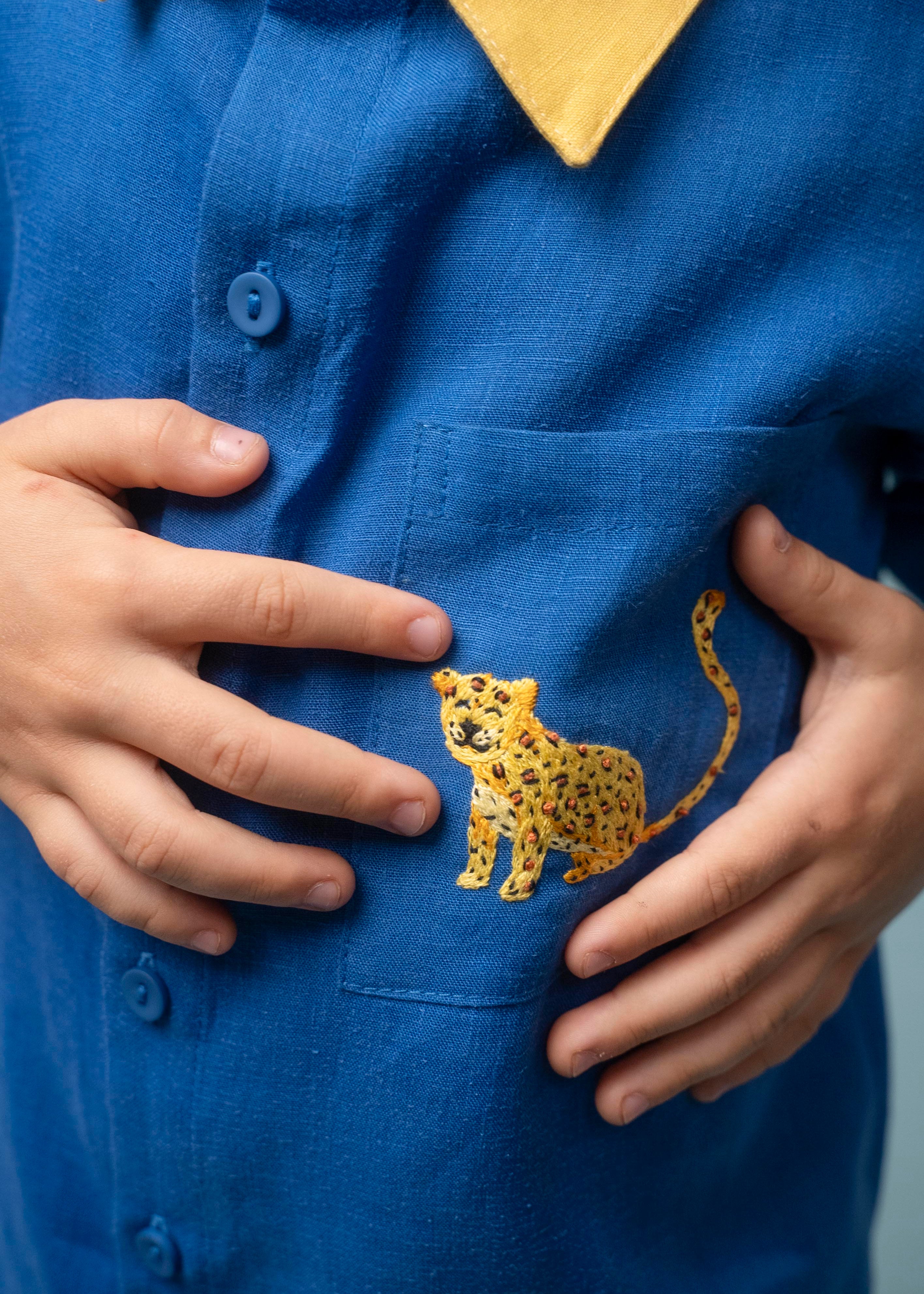 Camisa infantil azul em linho puro com gola amarela e bordado à mão de onça no bolso, usada por criança