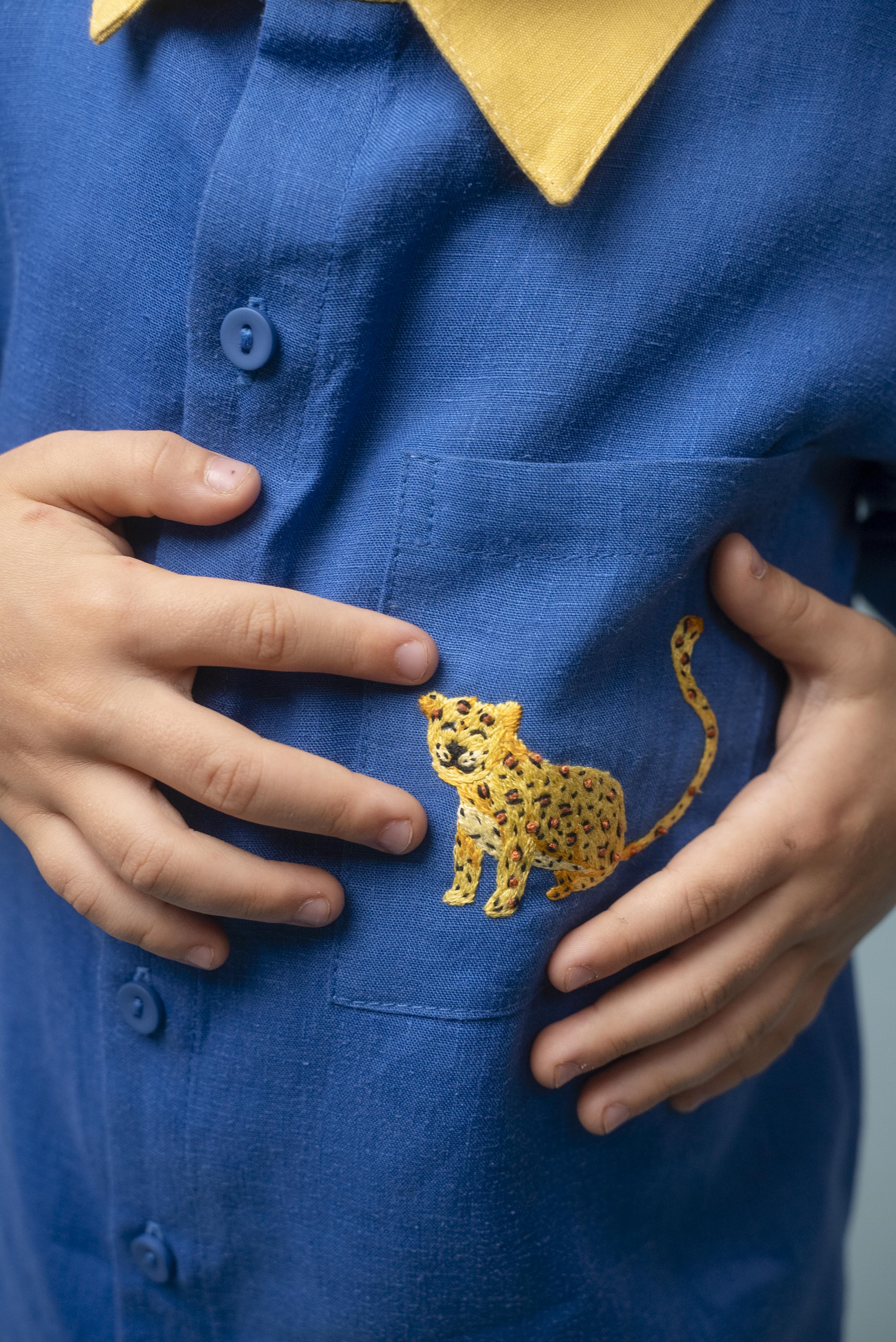 Camisa infantil azul em linho puro com gola amarela e bordado à mão de onça no bolso, usada por criança