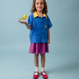 Menina usando camisa infantil azul e amarela em linho puro com bordado de onça, combinada com saia roxa