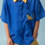 Menino usando camisa infantil de linho azul com gola amarela e bordado à mão de onça no bolso, combinada com shorts amarelo, visto em detalhe