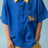 Menino usando camisa infantil de linho azul com gola amarela e bordado à mão de onça no bolso, combinada com shorts amarelo, visto em detalhe
