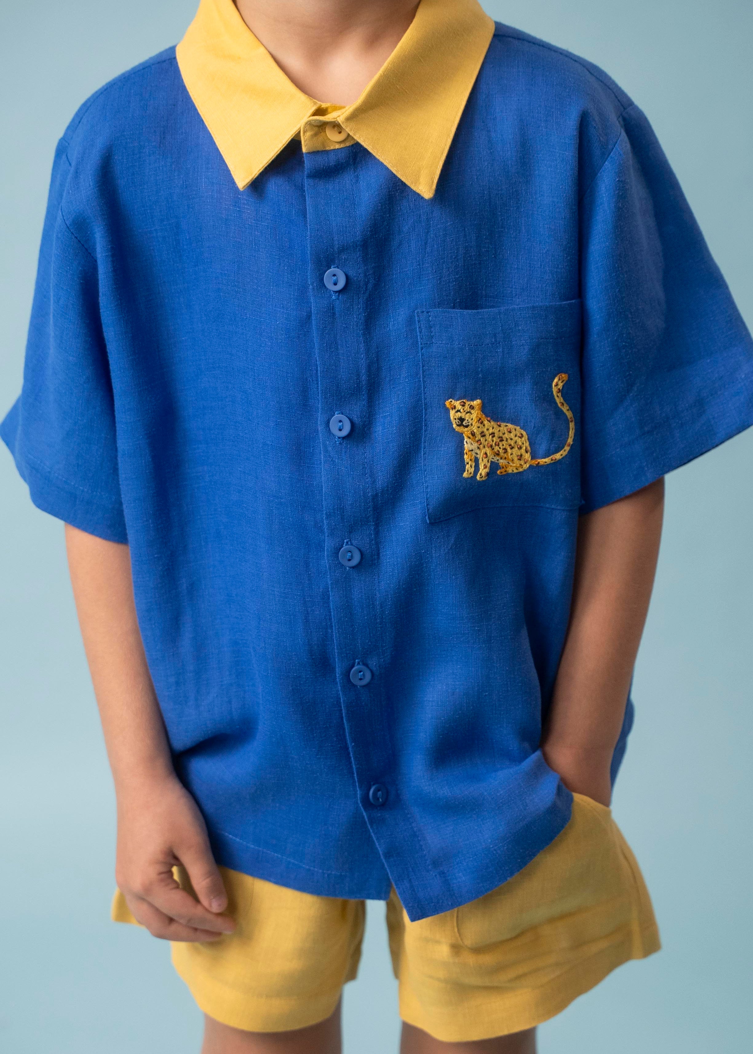 Menino usando camisa infantil de linho azul com gola amarela e bordado à mão de onça no bolso, combinada com shorts amarelo, visto em detalhe