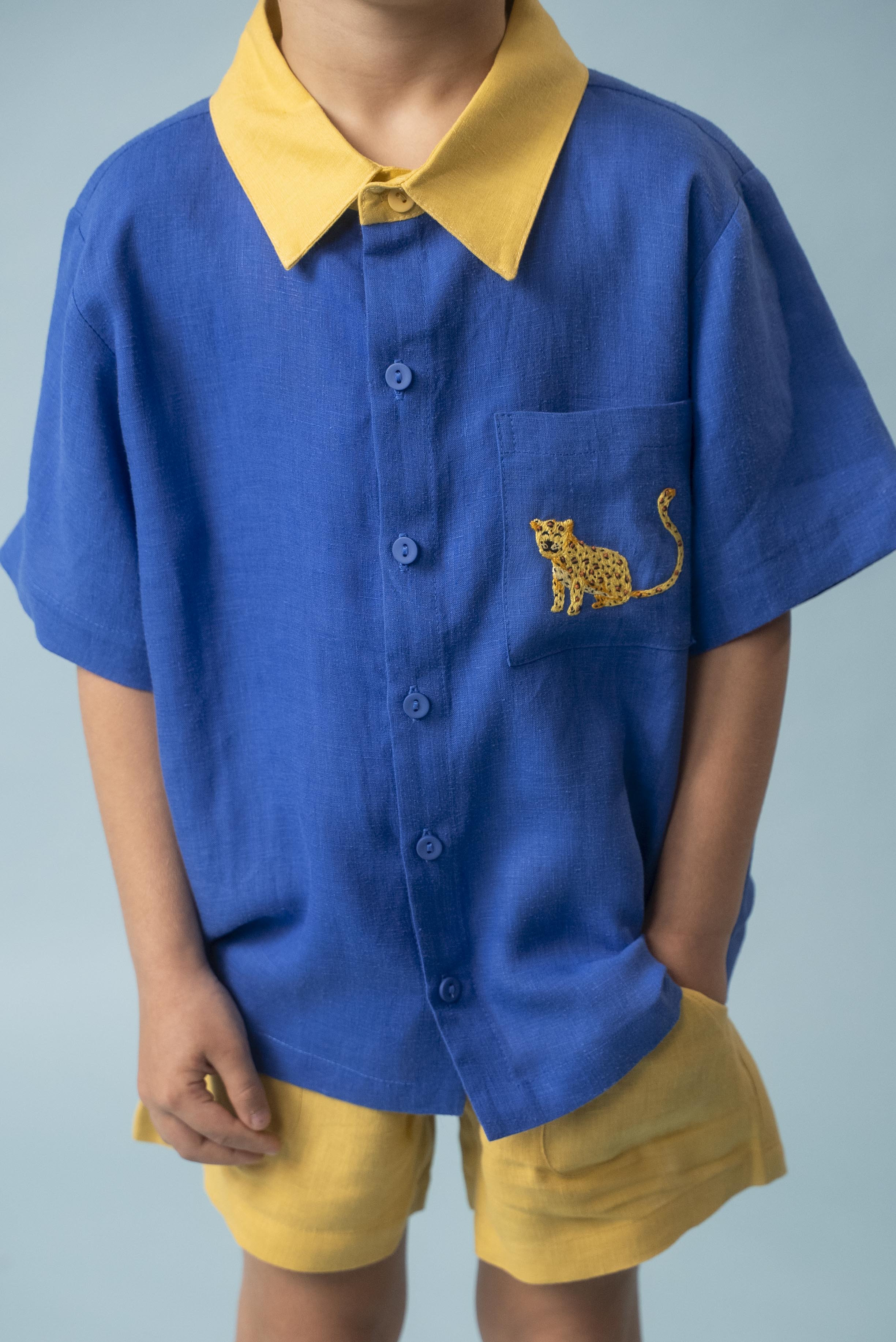 Menino usando camisa infantil de linho azul com gola amarela e bordado à mão de onça no bolso, combinada com shorts amarelo, visto em detalhe